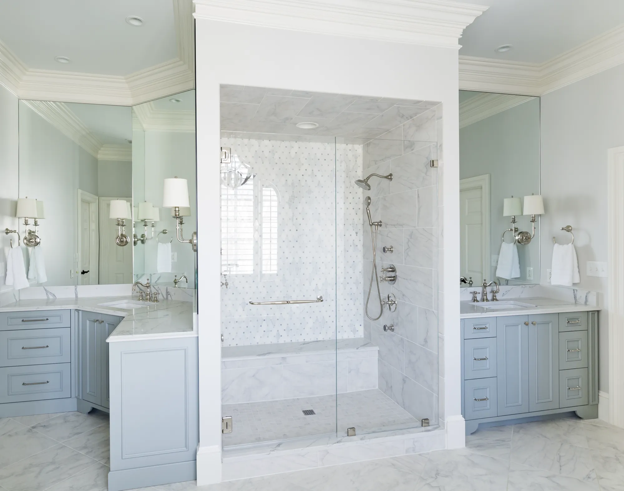 A bright bathroom featuring a central marble walk-in shower and two mirrored light gray vanities with white countertops and chrome fixtures.