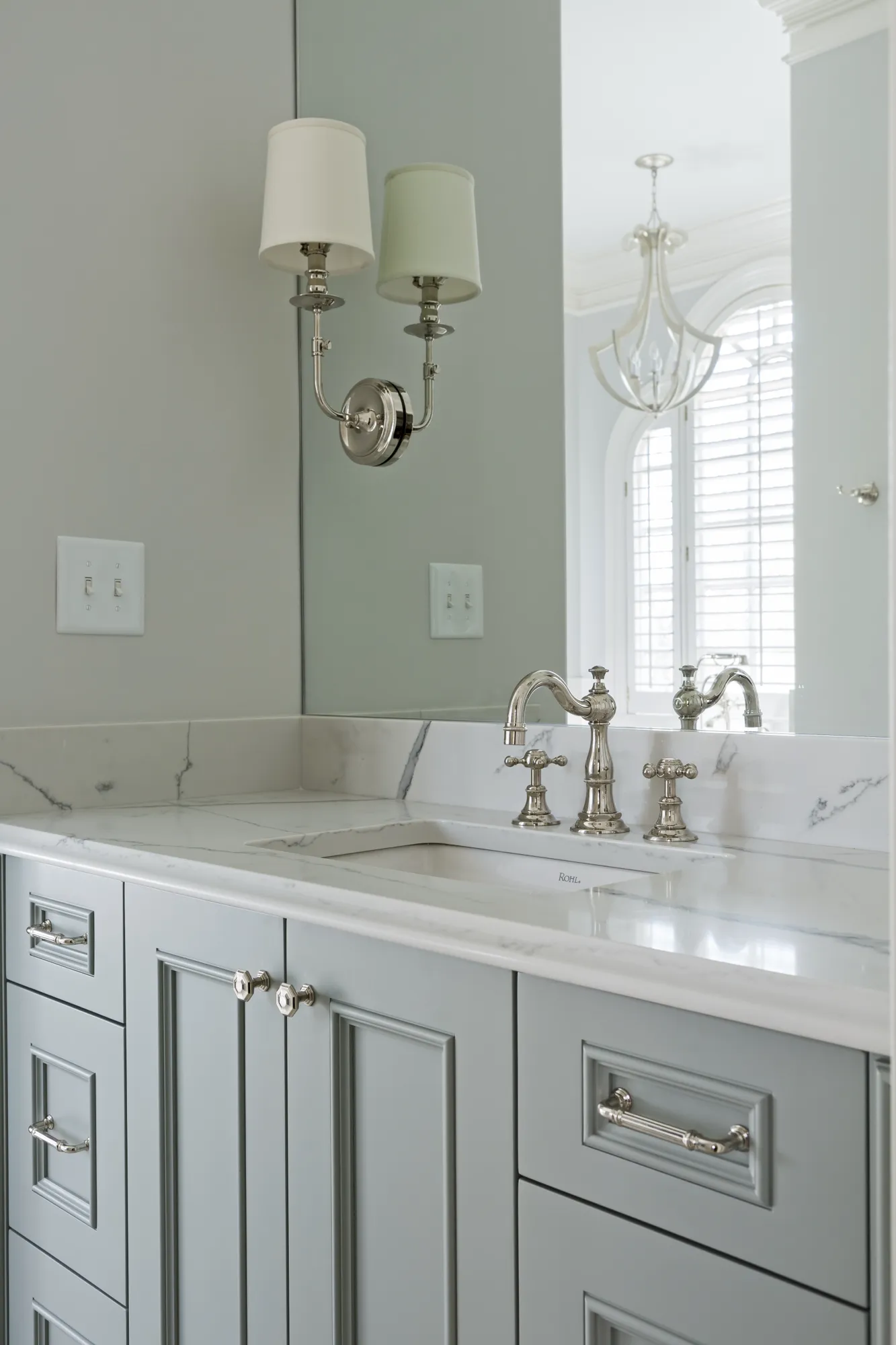 An elegant bathroom vanity featuring light gray paneled cabinets, a marble countertop, and polished nickel fixtures. The mirror reflects an arched window with white shutters.
