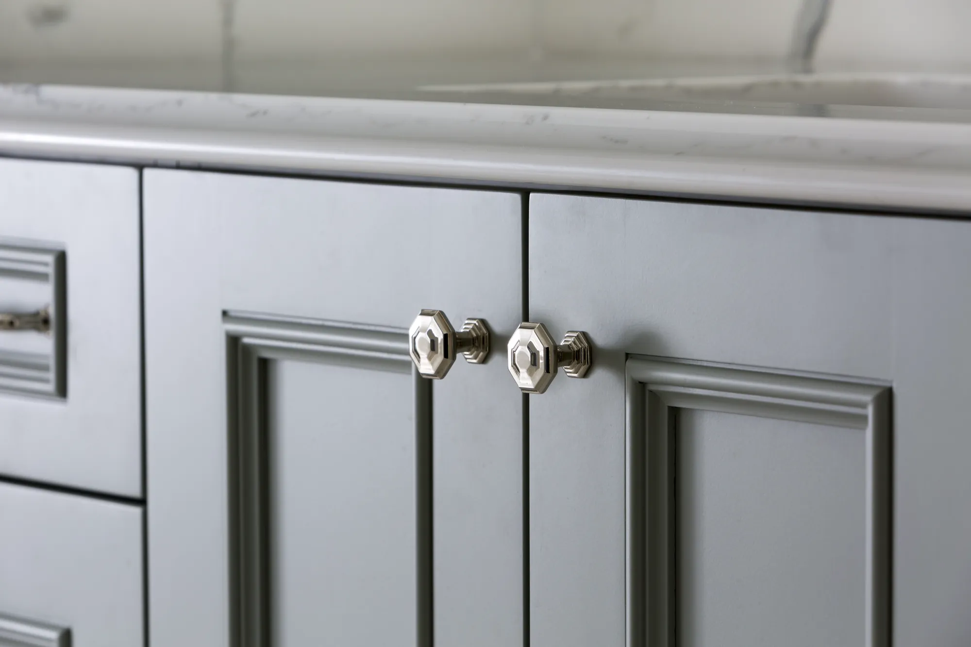 Recessed panel light gray cabinet doors featuring polished octagonal chrome knobs sit beneath a white marble countertop.