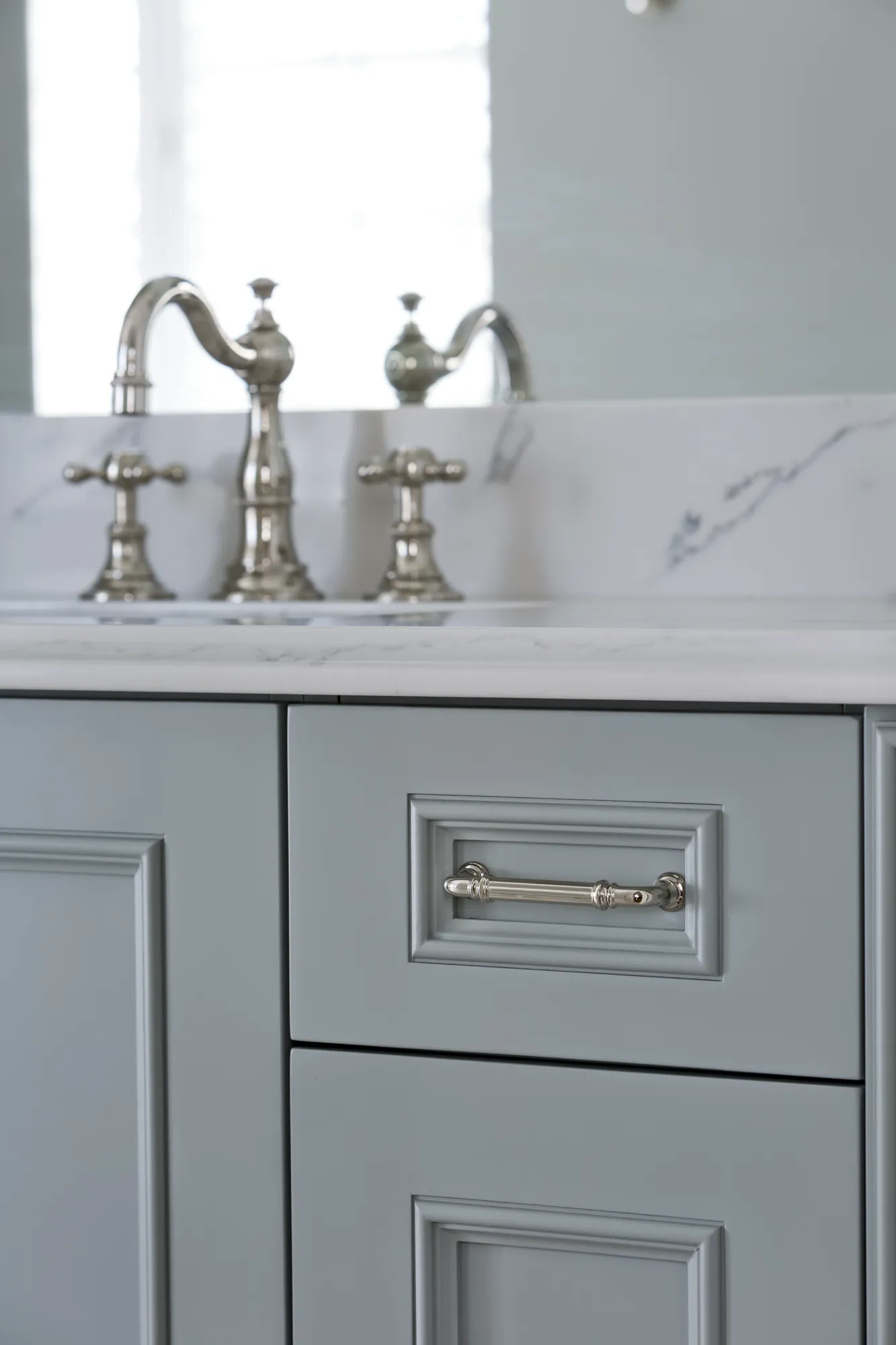 A blue-gray paneled vanity cabinet with a polished nickel handle is topped by a white marble counter and a silver three-piece faucet.