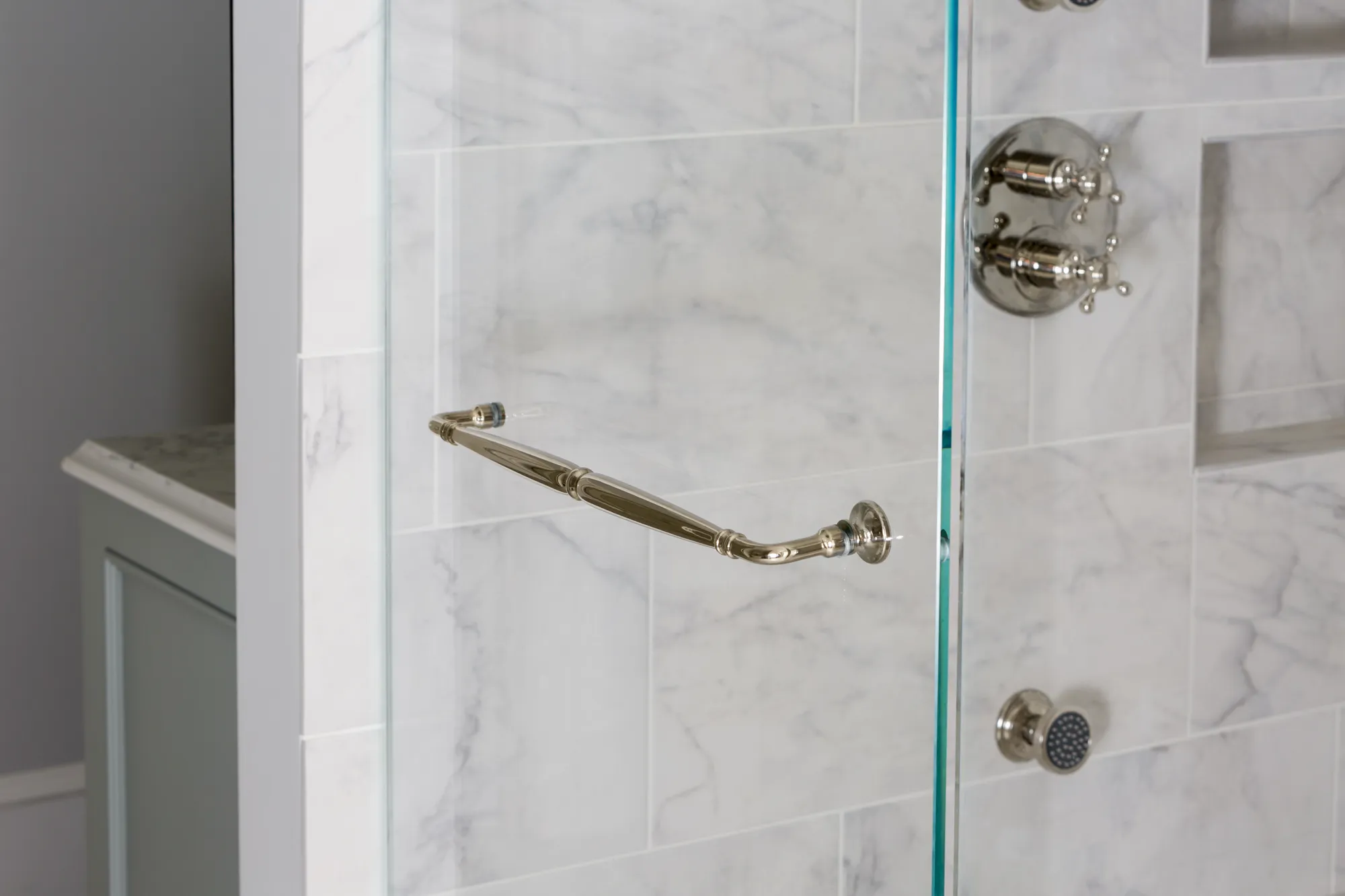 A clear glass shower door featuring a bright polished handle, set against a wall tiled with luxurious white rectangular marble. Fixtures and controls are visible inside.