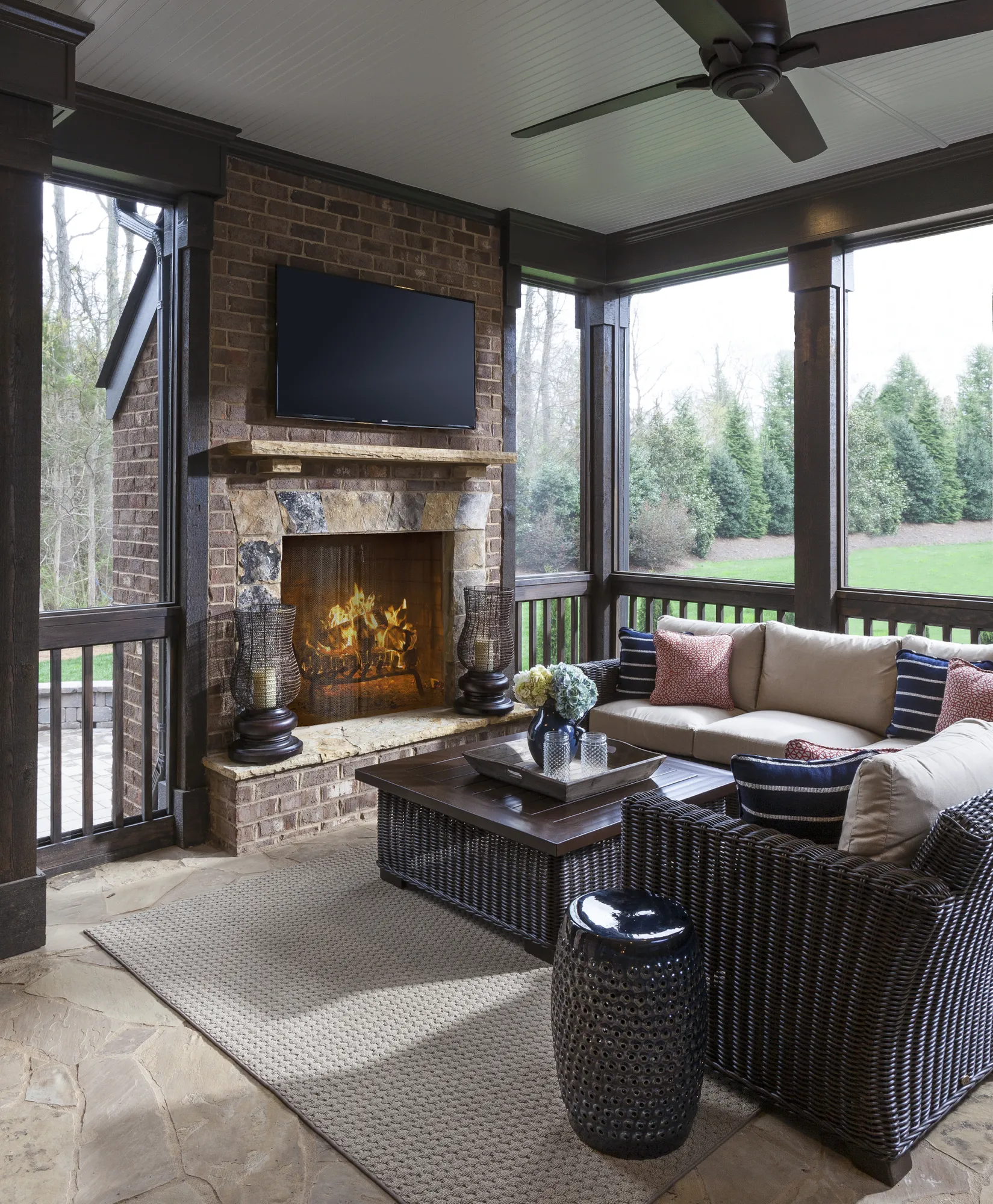 A high-end screened porch with dark wicker seating and a cozy stone fireplace, complete with a mounted TV. The room features stone flooring and an outdoor rug.