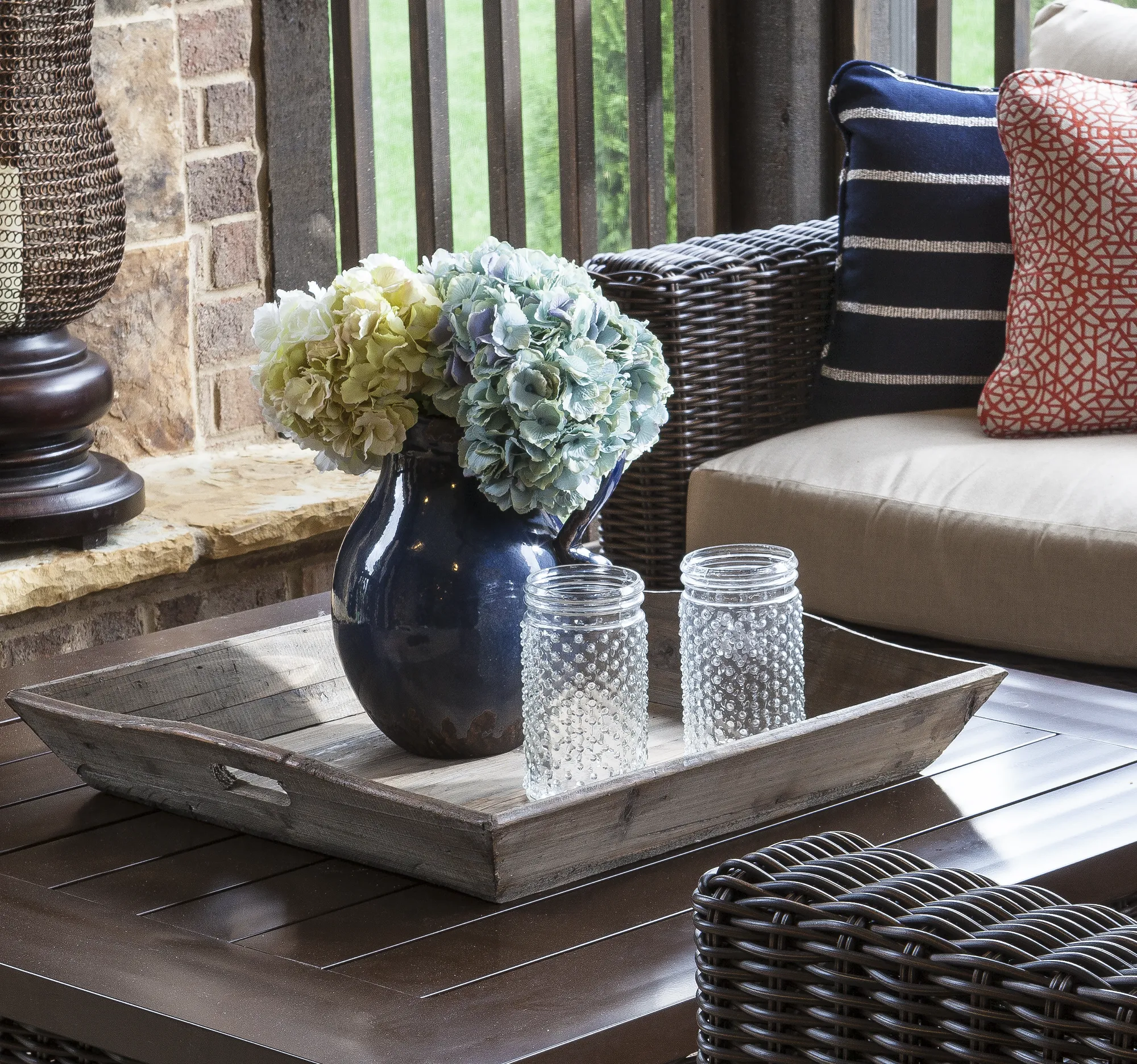 A rustic wooden tray on a dark outdoor table holds a blue vase of pale hydrangeas and two textured glass jars. Dark wicker furniture, tan cushions, and patterned pillows are visible.