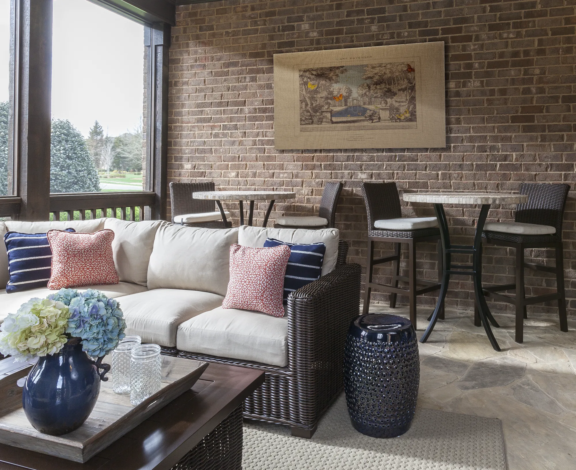A comfortable screened porch with a large wicker sectional, decorative pillows, and two high-top bar tables set against a dark brick accent wall.