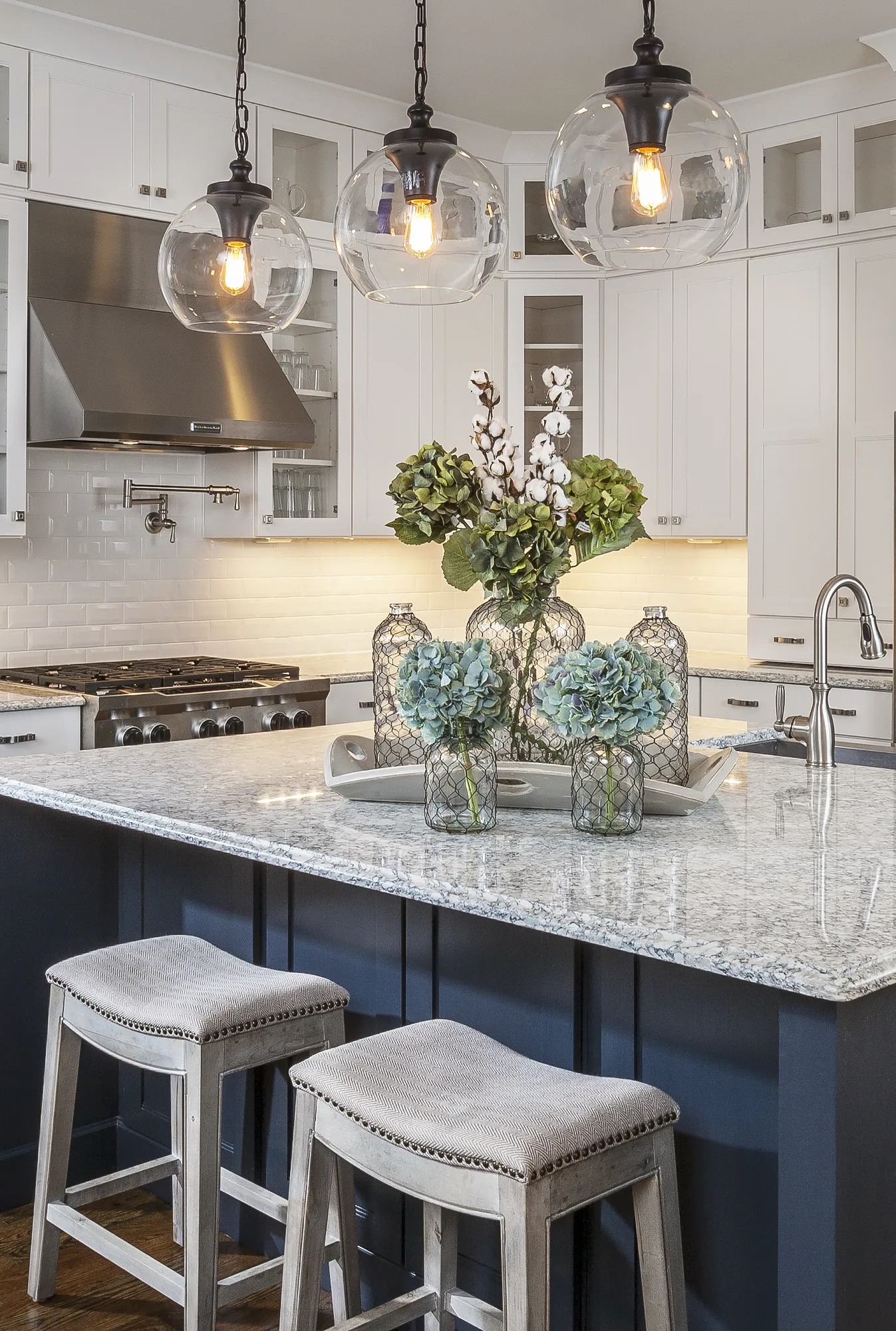 A clean, modern kitchen features a granite island with two stools and a dark blue base. Three globe pendant lights hang over a centerpiece of hydrangeas.
