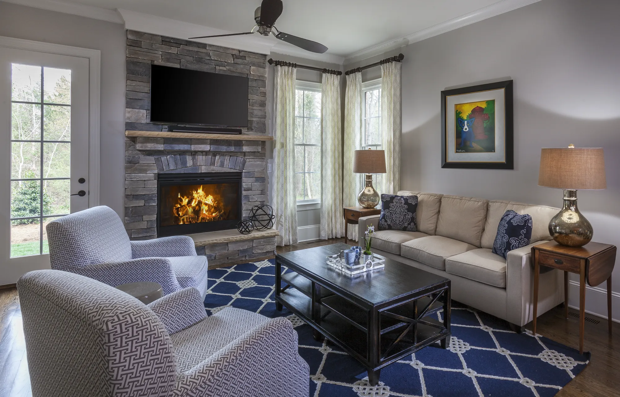 A cozy living room with a lit stone fireplace and a TV mounted above. The seating includes a tan sofa and two patterned chairs set around a coffee table on a vibrant blue geometric rug.