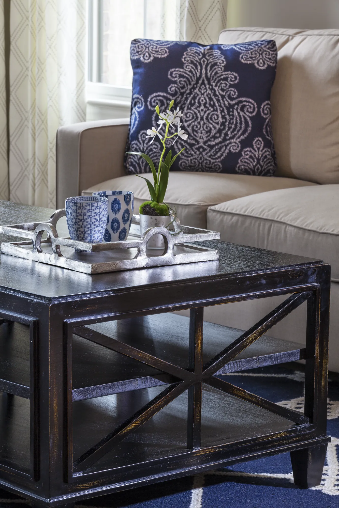 A close-up of a dark coffee table holding a silver tray with two blue patterned mugs and a small white orchid plant, next to a beige couch.