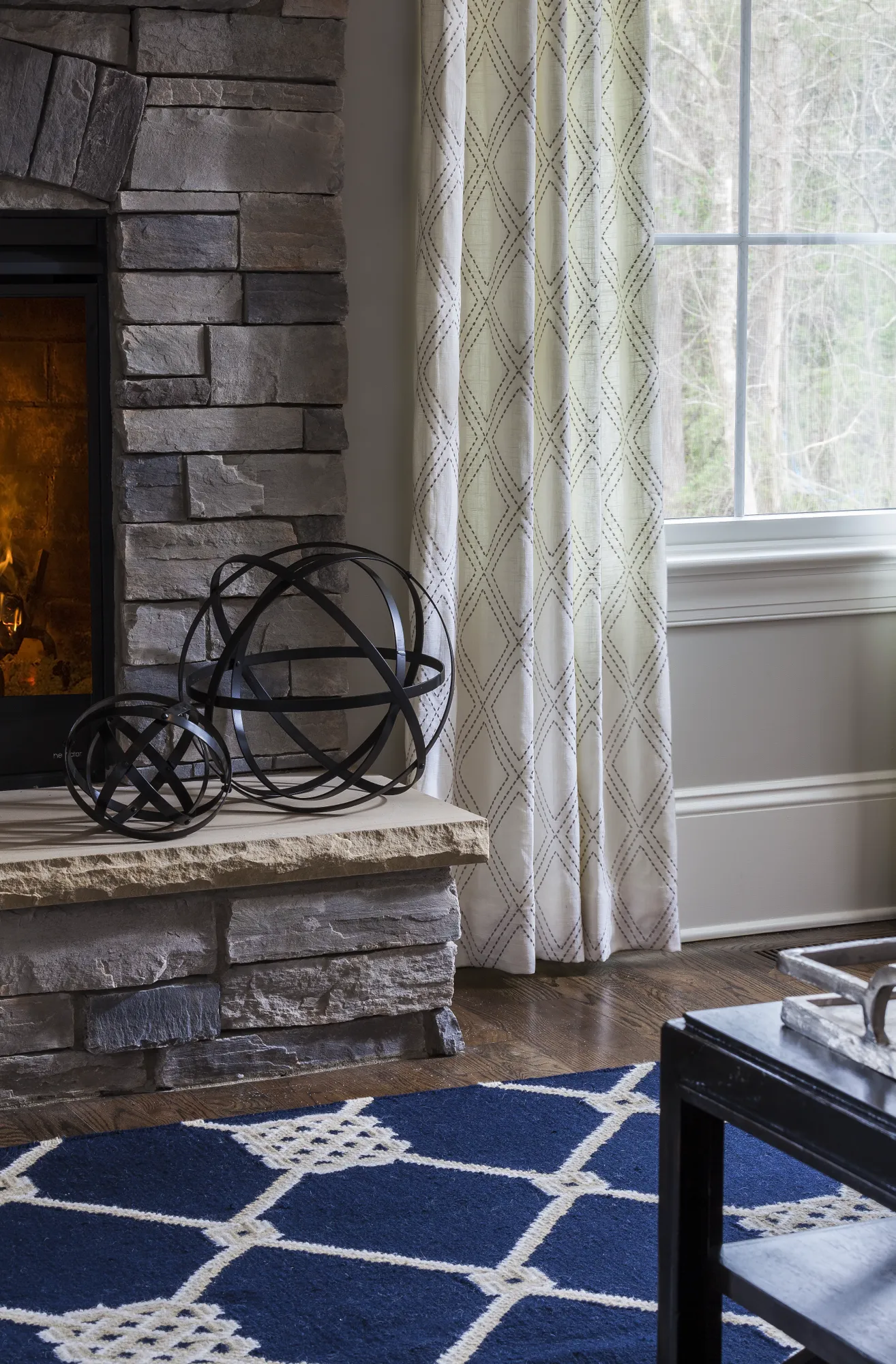 A stone fireplace flanked by sheer patterned curtains and a geometric navy blue rug. Two dark metal spheres decorate the beige stone hearth.