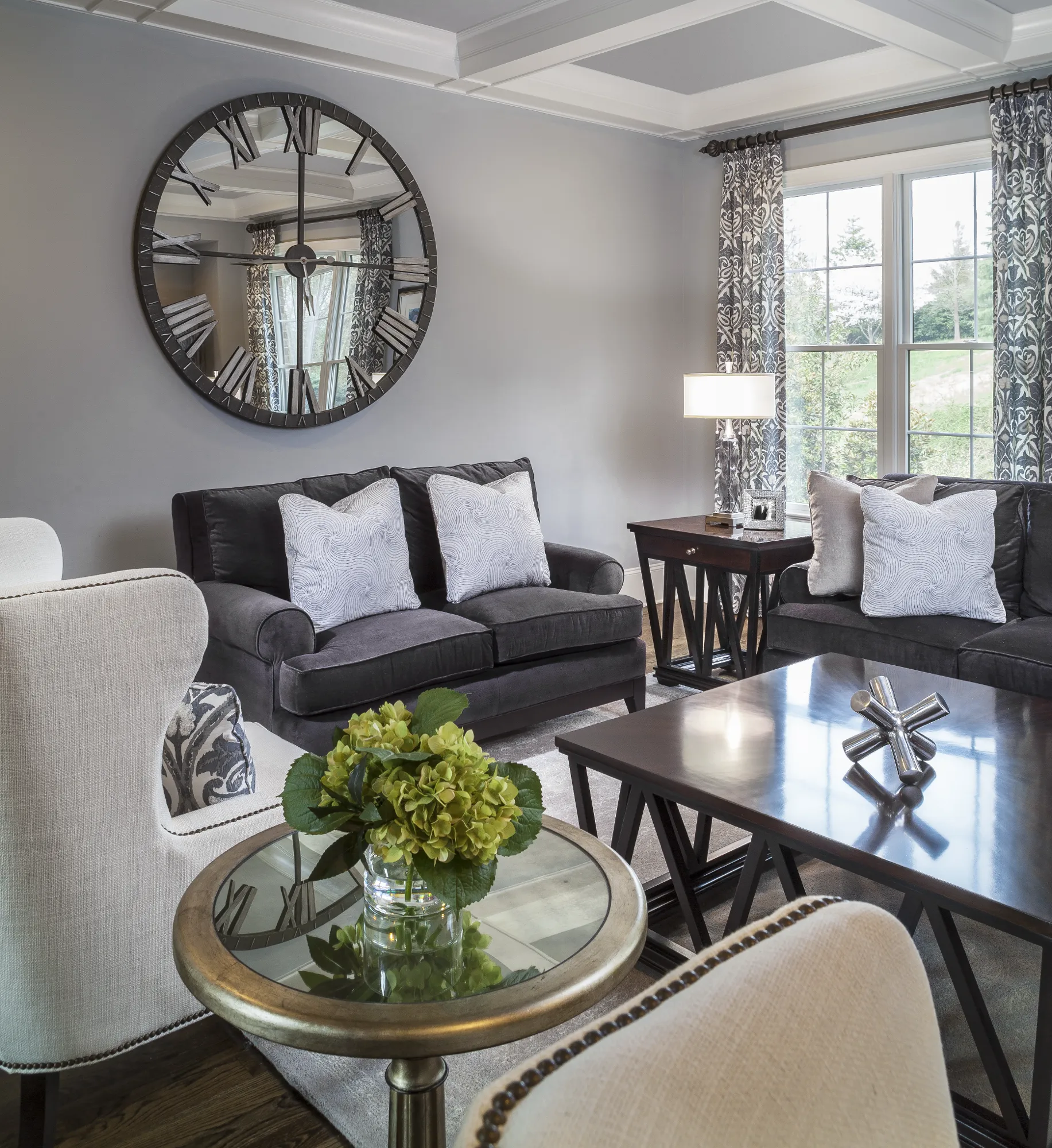 A sophisticated living area with dark sofas, light upholstered chairs, and a large mirrored Roman numeral wall clock. A coffered ceiling and patterned curtains complete the refined design.