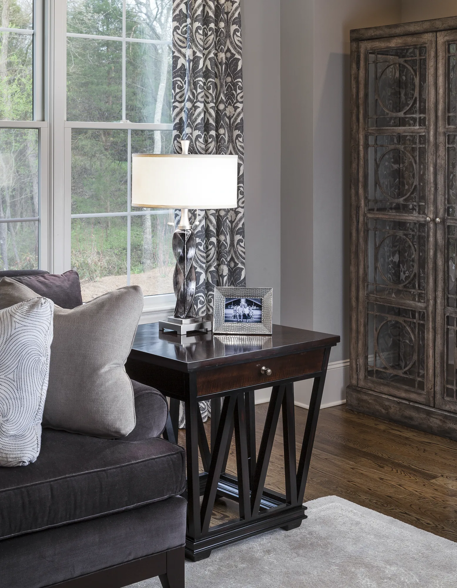 An elegant living room corner featuring a dark wooden end table next to a plush sofa with throw pillows. A lamp, a framed photo, and a dark patterned curtain are visible against a large window.