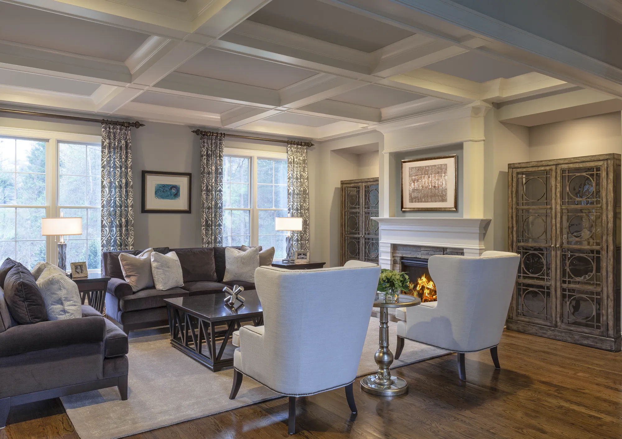 An elegant and cozy living room featuring a white coffered ceiling, a dark velvet sectional, two light armchairs, and a fireplace with a lit fire.