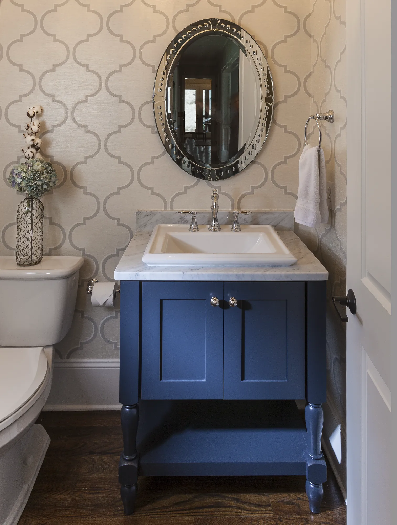A stylish powder room featuring a navy blue vanity, a marble sink counter, and an ornate oval mirror against light quatrefoil pattern wallpaper.