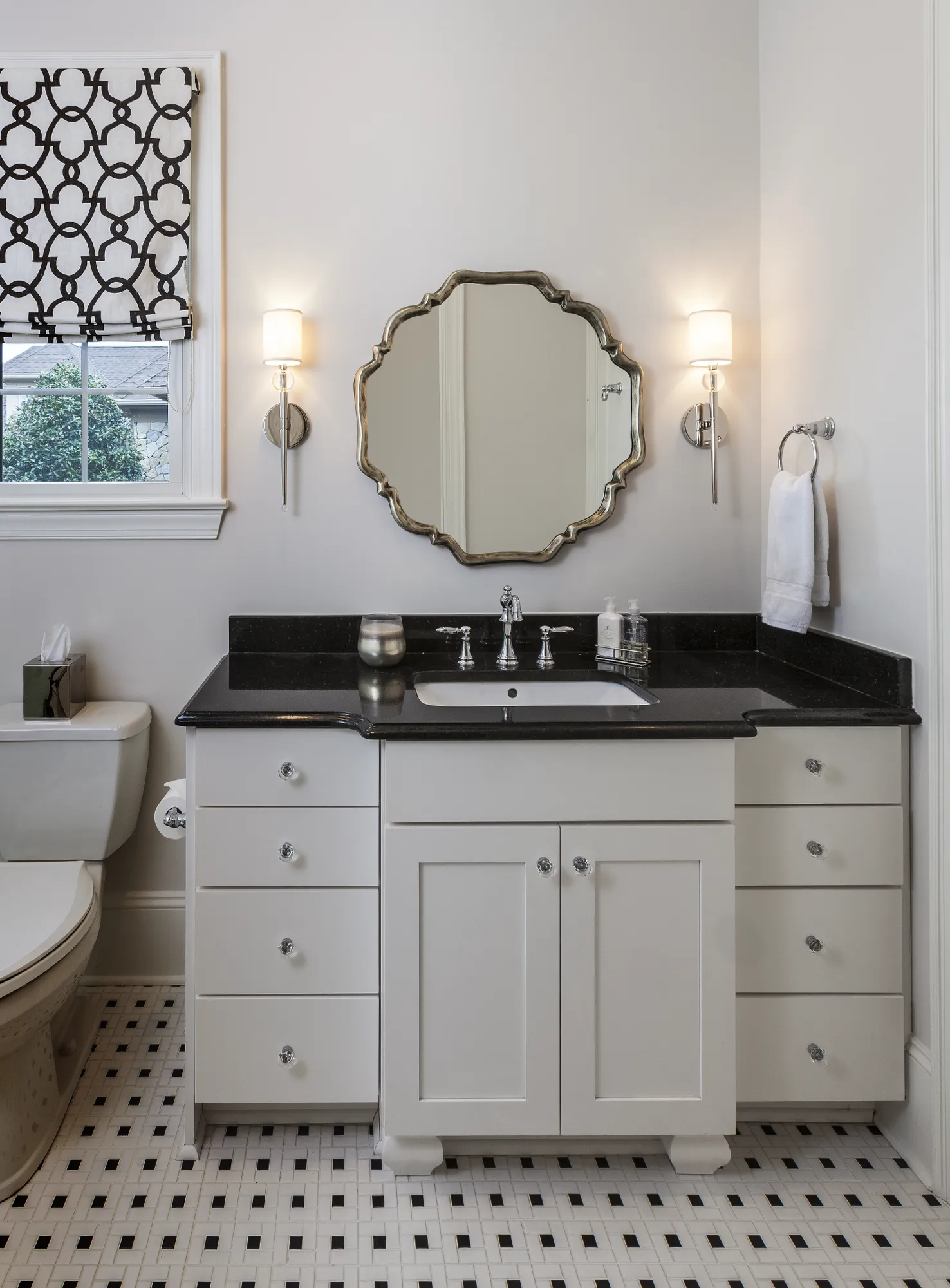 A white vanity with a black granite counter stands beneath a scalloped mirror and two sconces in a bathroom with black and white mosaic tile.