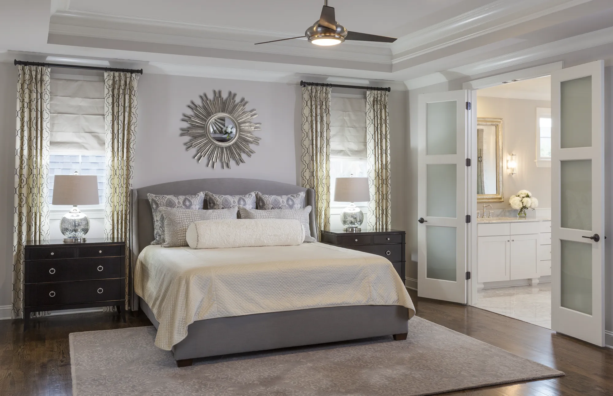 A luxurious bedroom featuring a gray upholstered bed and symmetrical nightstands. Double glass doors reveal a bright, white en-suite bathroom with a vanity and marble counter.