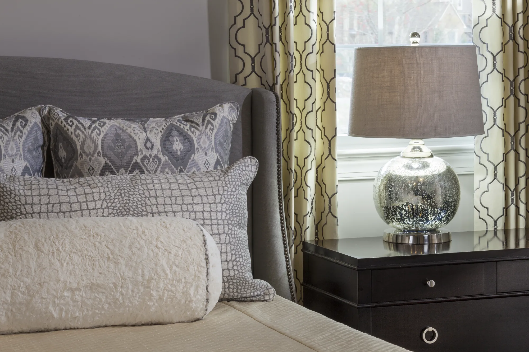 A luxurious bedroom featuring a gray upholstered headboard, decorative pillows, and a dark nightstand with a mercury glass lamp. Patterned curtains frame the window.