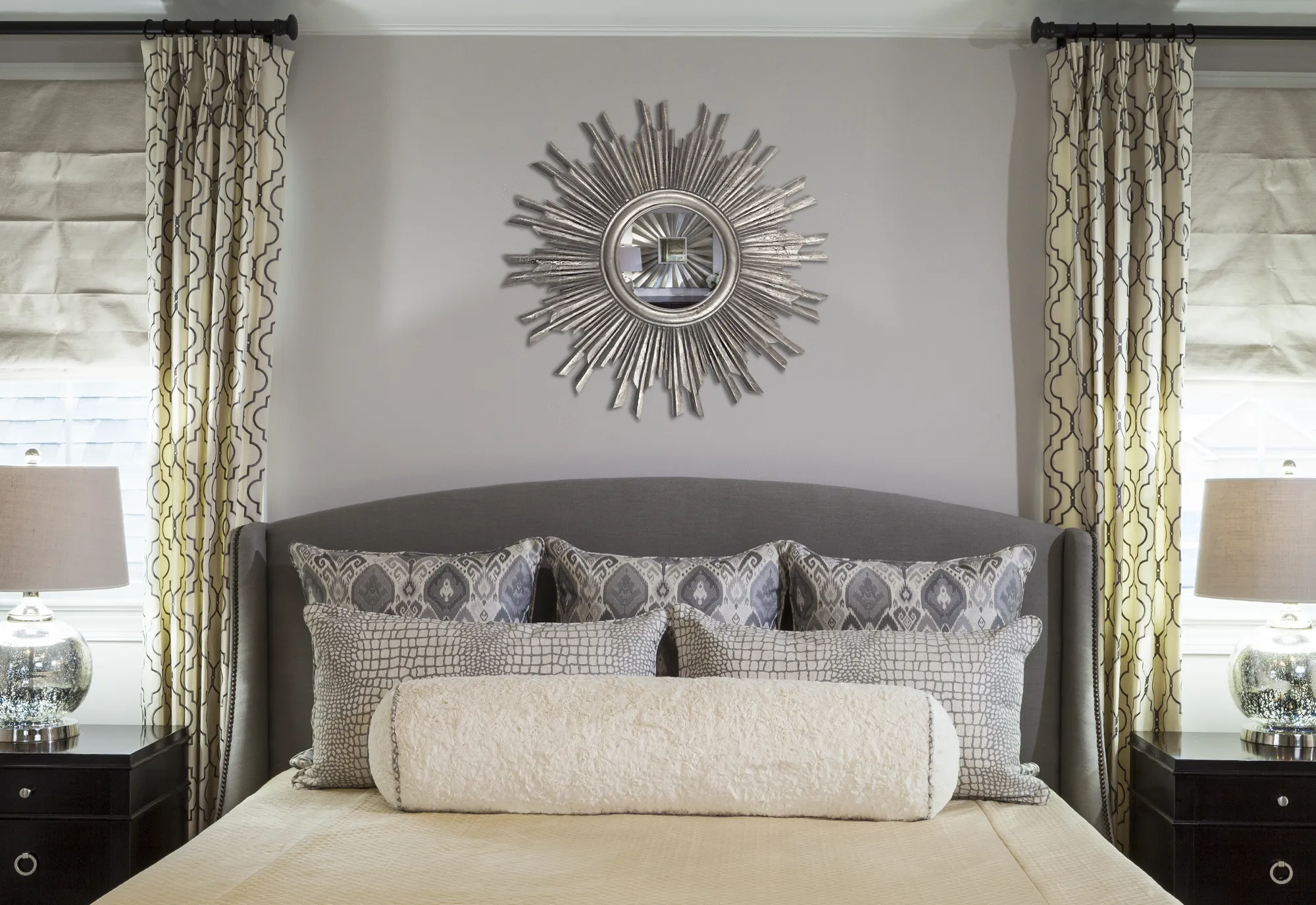 A sophisticated bedroom featuring a gray upholstered headboard with patterned pillows. A silver sunburst mirror hangs above, flanked by two dark nightstands and matching lamps.