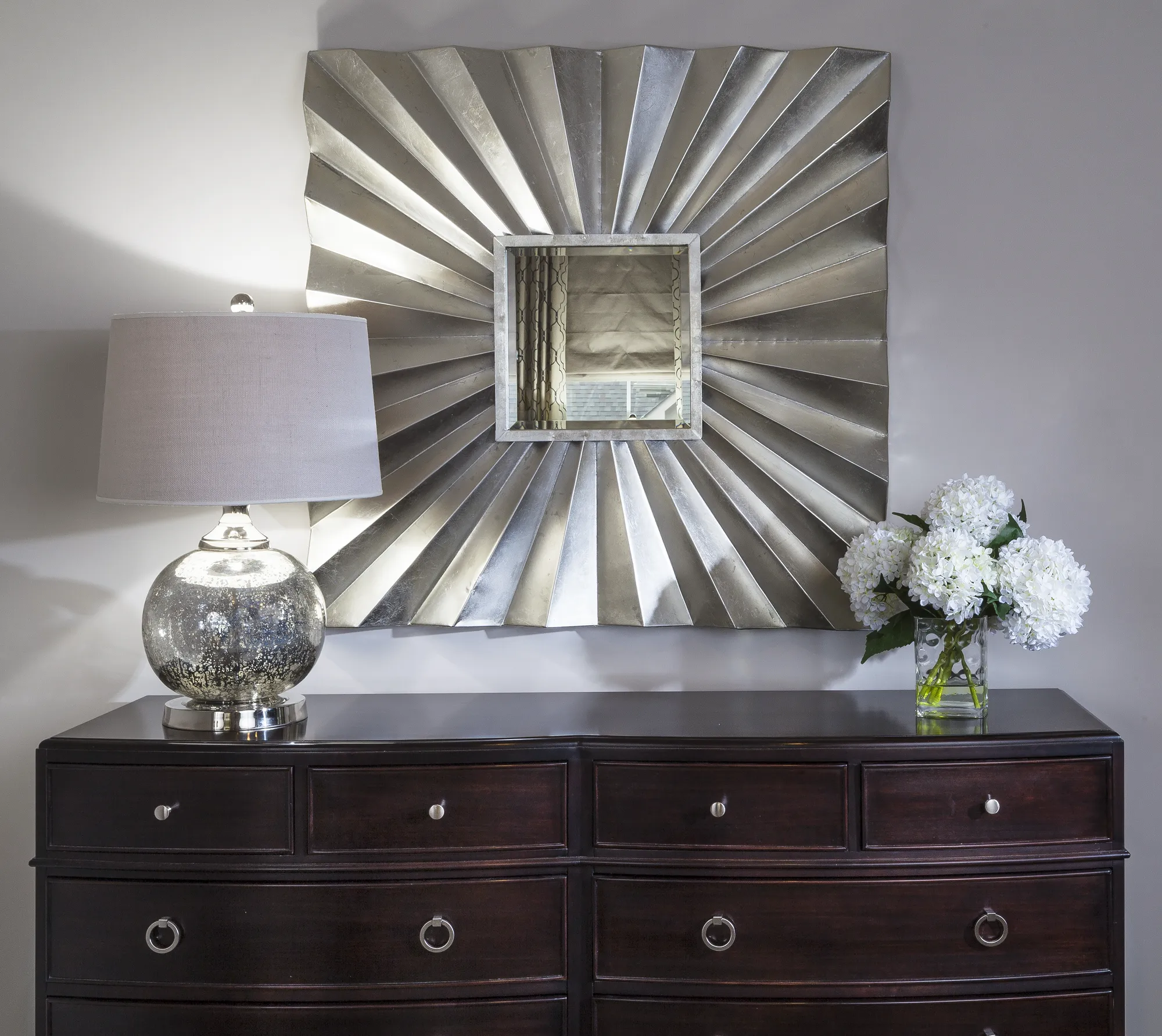 An elegant setting features a dark wood chest beneath a large, silver sunburst mirror, accented by a lamp and white hydrangeas.