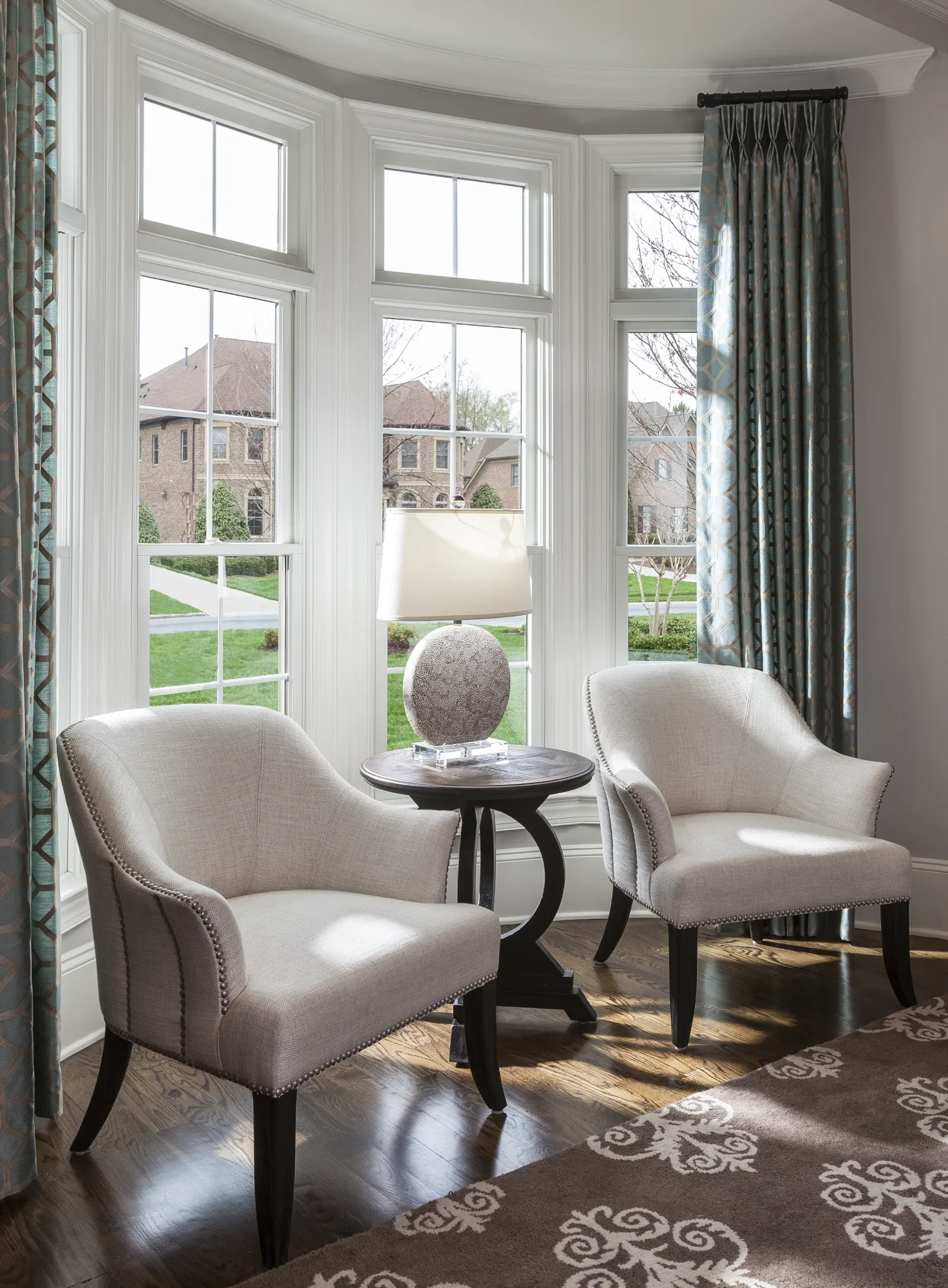 Two elegant, light beige chairs with nailhead trim and a lamp table sit in a sunny bay window nook with patterned curtains.