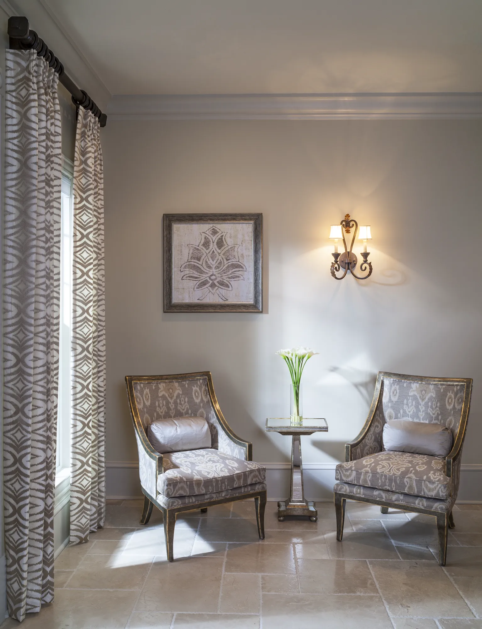 Two elegant, upholstered armchairs flank a side table with white flowers in a sophisticated room with patterned curtains, wall art, and an ornate sconce.