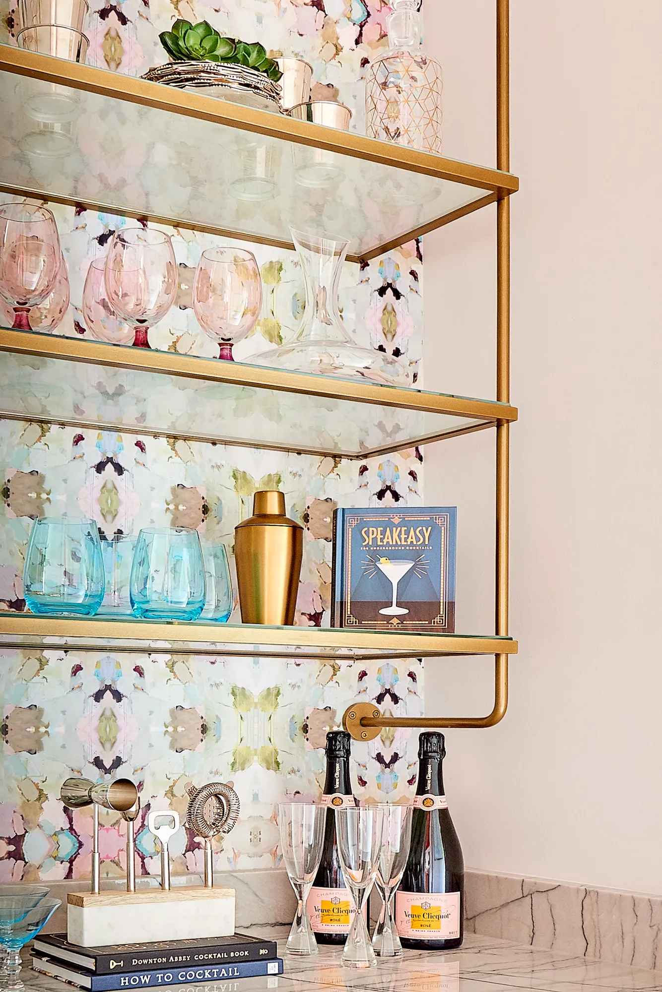 A sleek bar display with gold and glass shelving set against abstract, colorful wallpaper. The shelves hold various pink and blue glassware, a gold shaker, and cocktail tools and books.