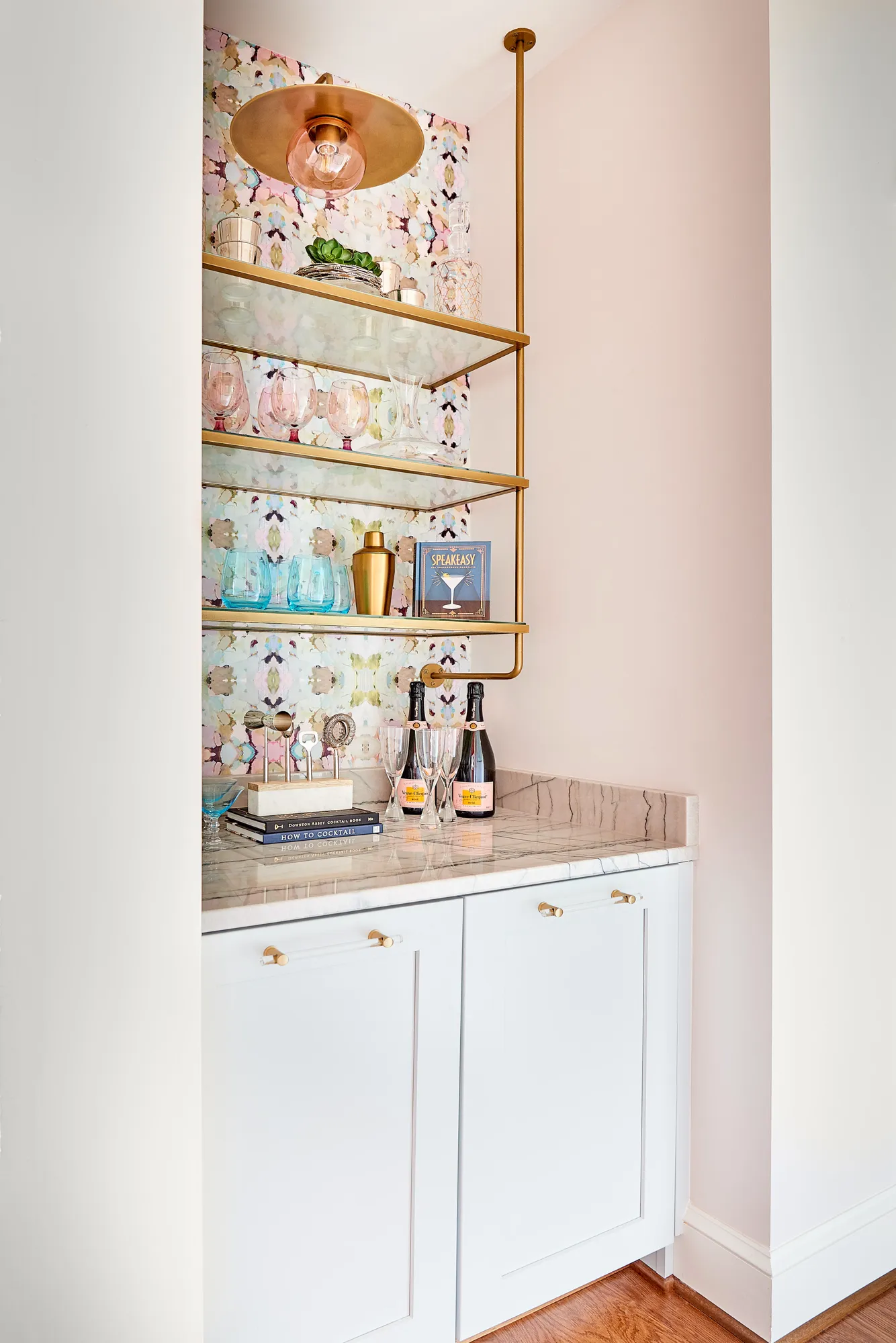 An elegant home bar with white cabinetry, a marble counter, and brass-framed glass shelves. The nook is lined with colorful, abstract wallpaper and stocked with glassware and champagne.