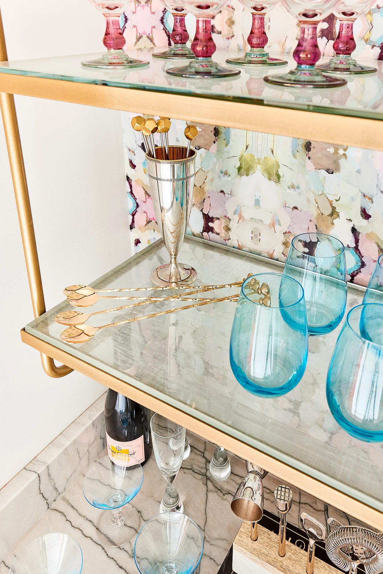 A chic gold and glass bar cart displays an array of colorful barware, including blue tumblers, pink stemmed glasses, and professional silver mixing tools.