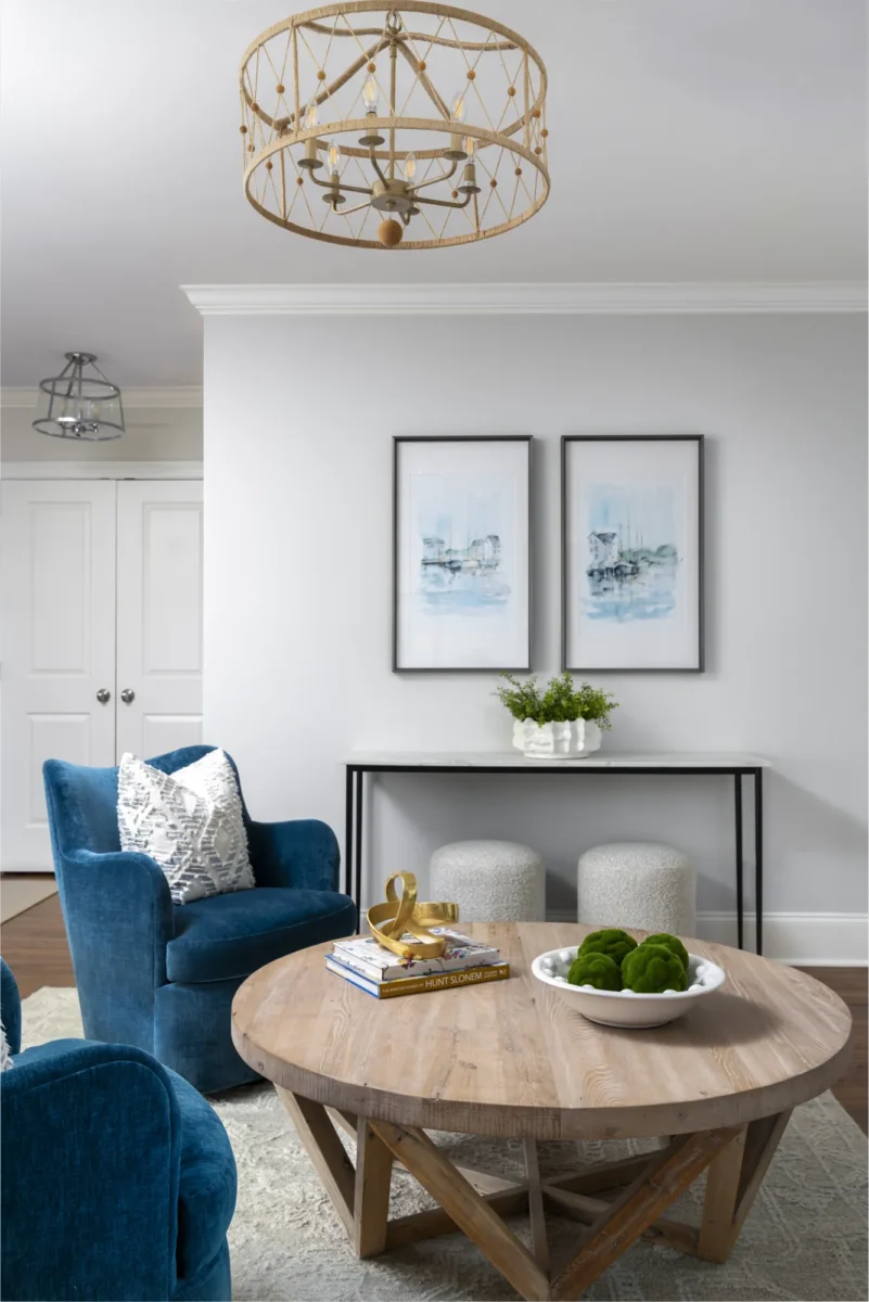 A bright living room showcasing coastal decor, including a weathered wood coffee table and blue velvet chairs. A rope chandelier illuminates the space above two nautical prints.