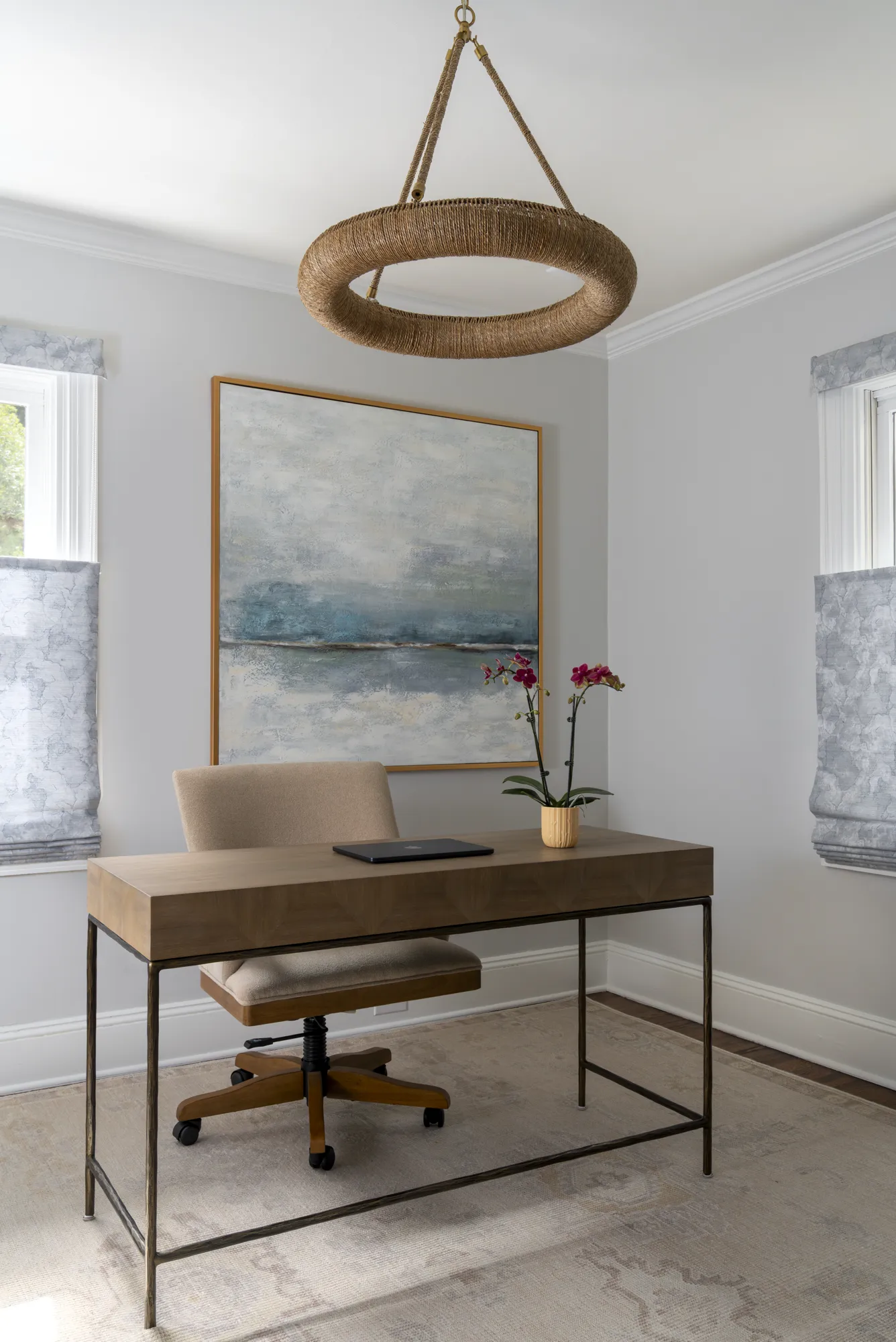 A clean home office with a wood desk and upholstered chair. Above the desk, a large abstract painting hangs beneath a unique rope-wrapped circular light fixture.