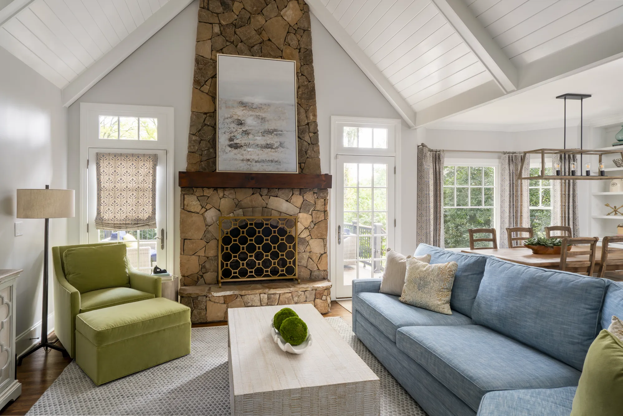 A great room features a floor-to-ceiling stone fireplace and vaulted shiplap ceiling. A blue sofa and green armchair frame the fireplace.