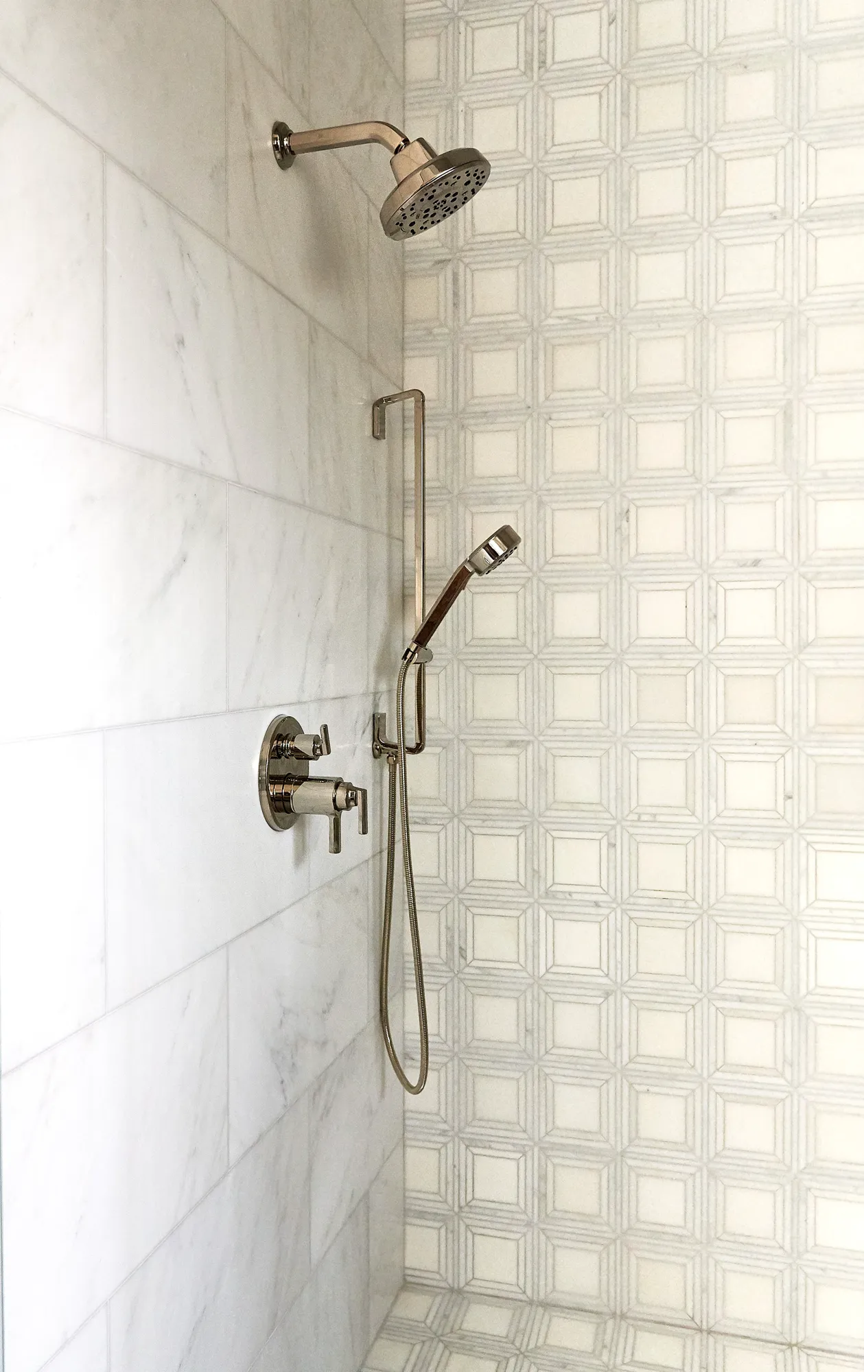 A luxury shower interior with polished chrome fixtures and two types of white marble tile: large-rectangular tiles and a geometric square mosaic wall.