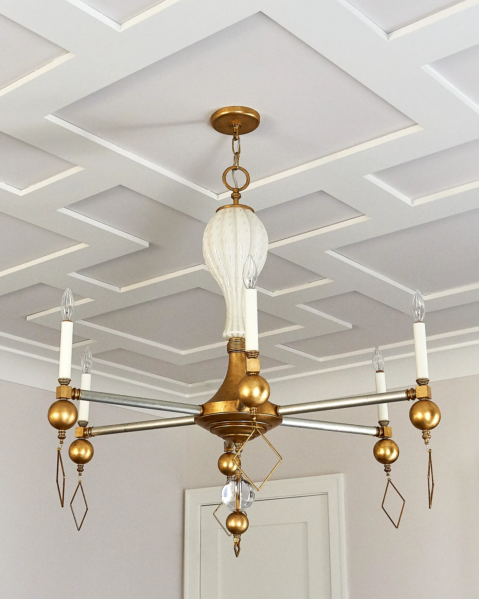 A decorative gold and silver chandelier with six candle lights and a ribbed white glass shade hangs from a white coffered ceiling.