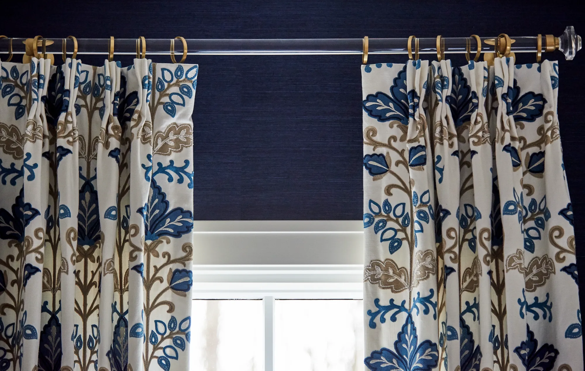 White curtains with a blue and taupe floral pattern hang on a clear rod with brass rings, set against a navy blue textured wall above a bright window.