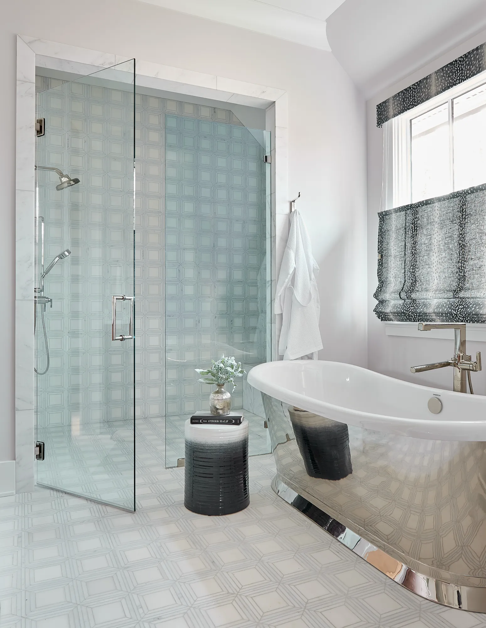 A bright, modern bathroom features a glass shower enclosure next to a freestanding white tub with a reflective silver base. The floor and shower wall have geometric patterned tiles.