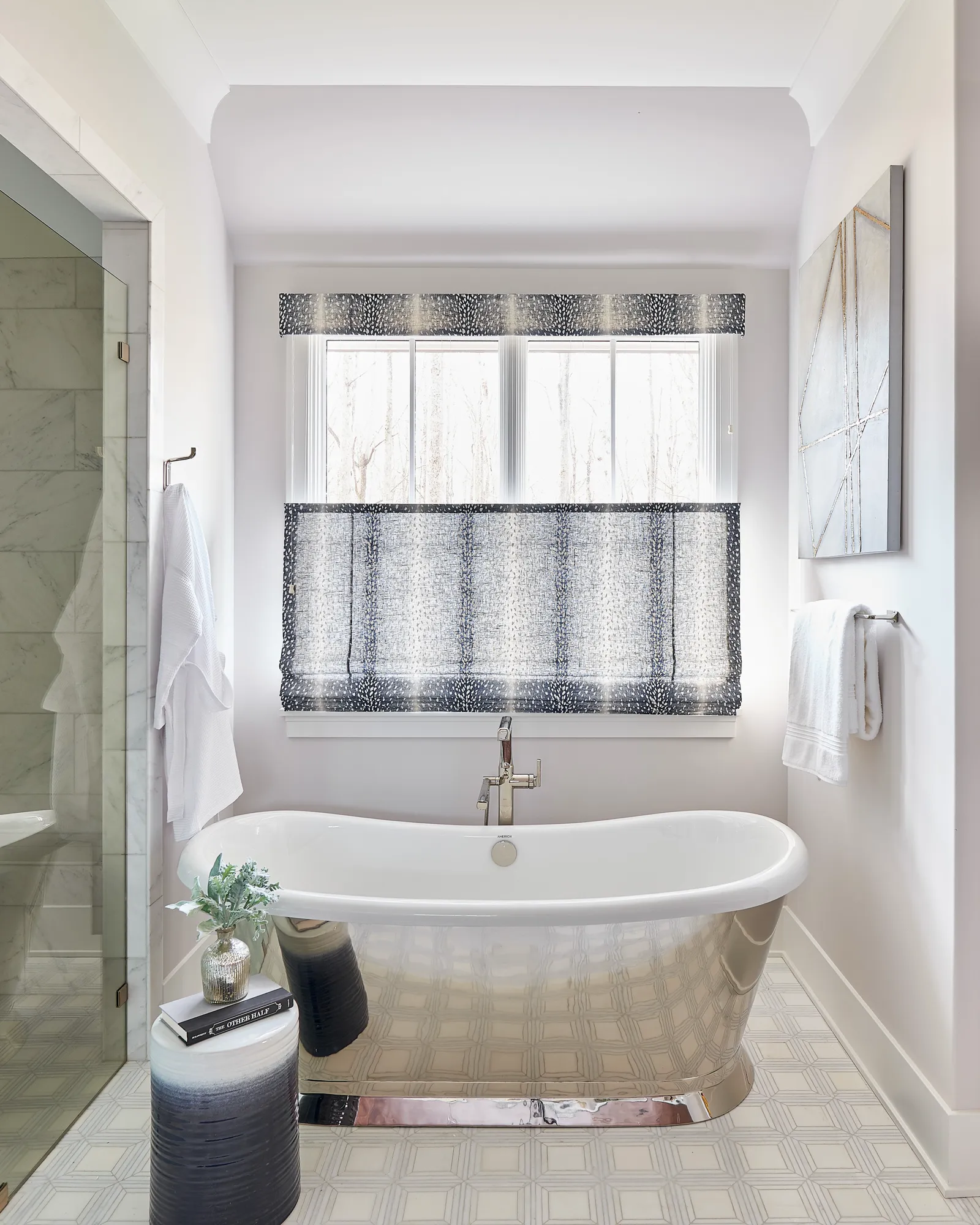 A modern, bright bathroom with a chrome-finished freestanding tub centered below a window with patterned Roman shades. Geometric floor tiles and a glass shower enclosure complete the design.