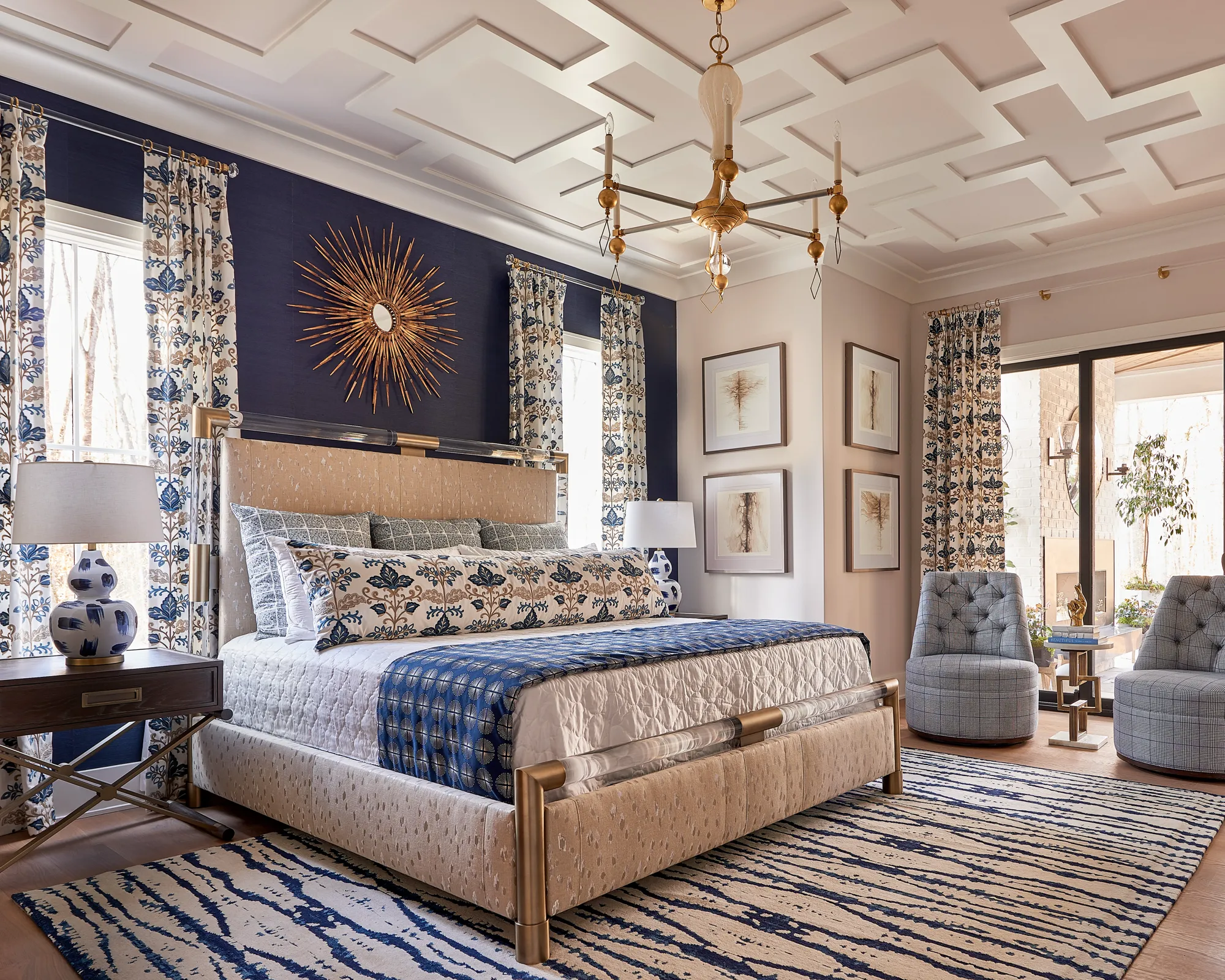 A luxurious bedroom with a tan upholstered bed and a navy feature wall accented by a gold sunburst mirror. A blue and white patterned rug anchors the space under a coffered ceiling.