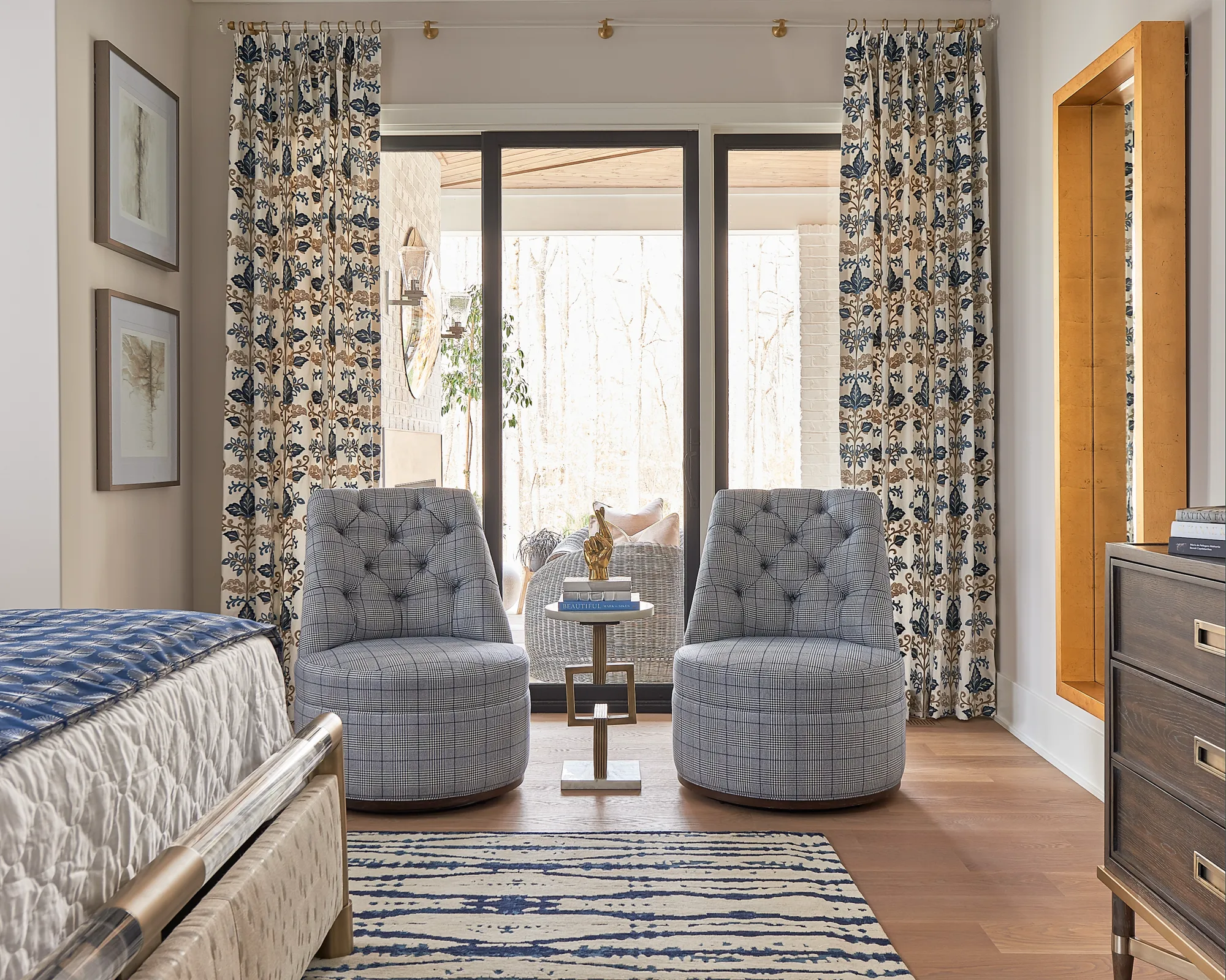 A sitting area features two round, tufted chairs in blue plaid, separated by a side table. Patterned drapes frame glass doors leading to a bright, wooded patio.