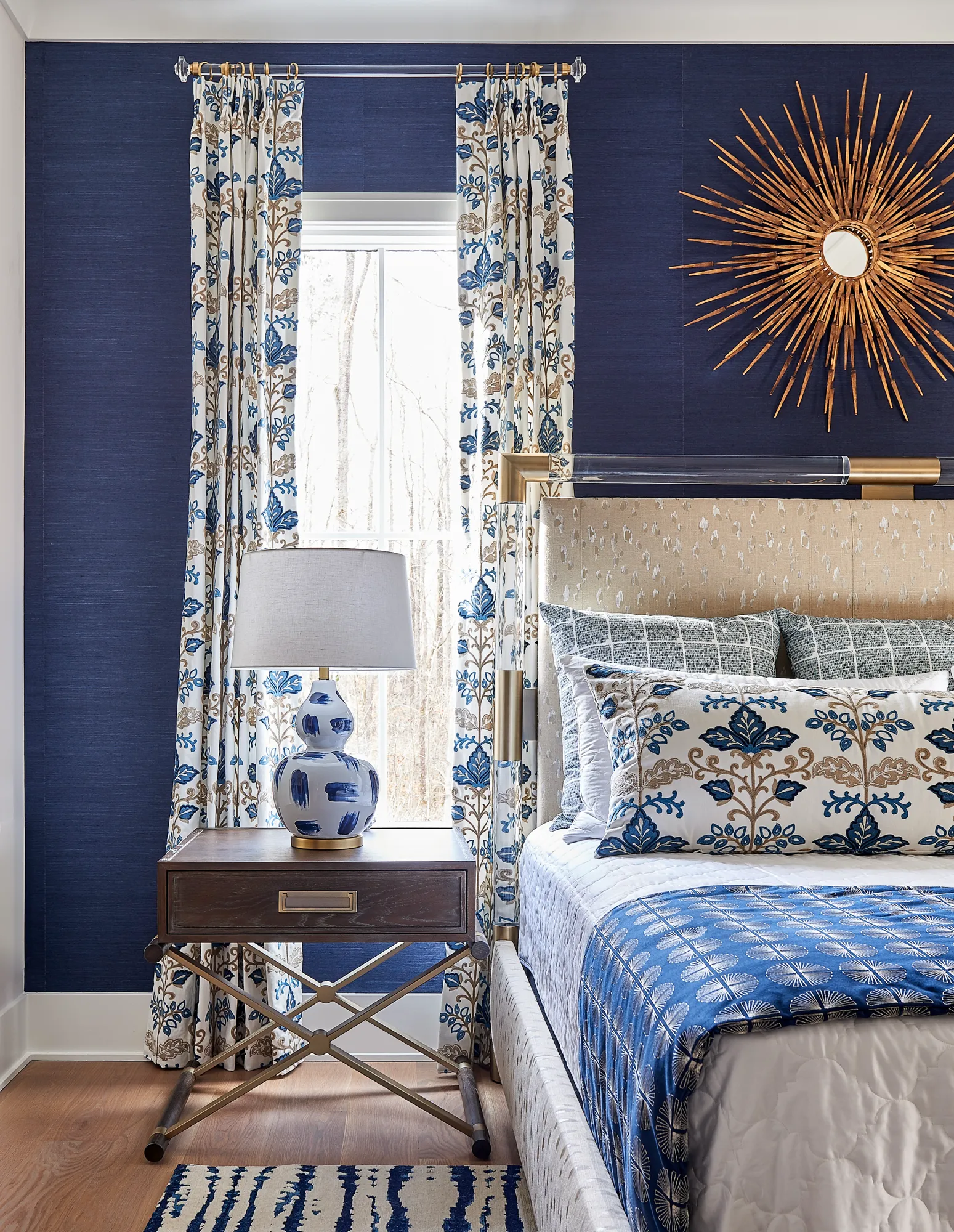 A stylish bedroom features navy blue walls, a beige upholstered headboard, and patterned blue and cream bedding. A sunburst mirror and matching curtains add dramatic accents.