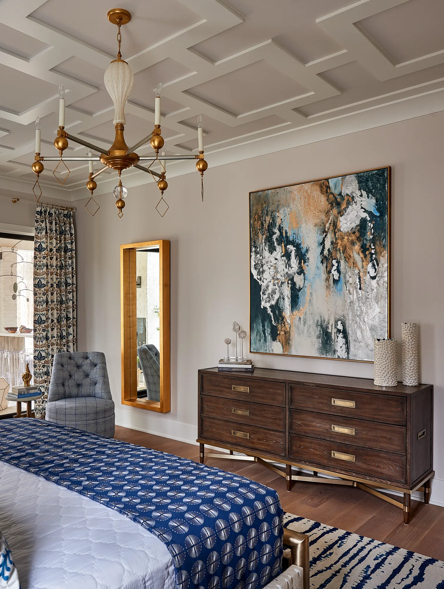 A sophisticated bedroom featuring a detailed geometric ceiling and a gold chandelier. Dark wood furniture and a large abstract painting complement the bold blue textiles.