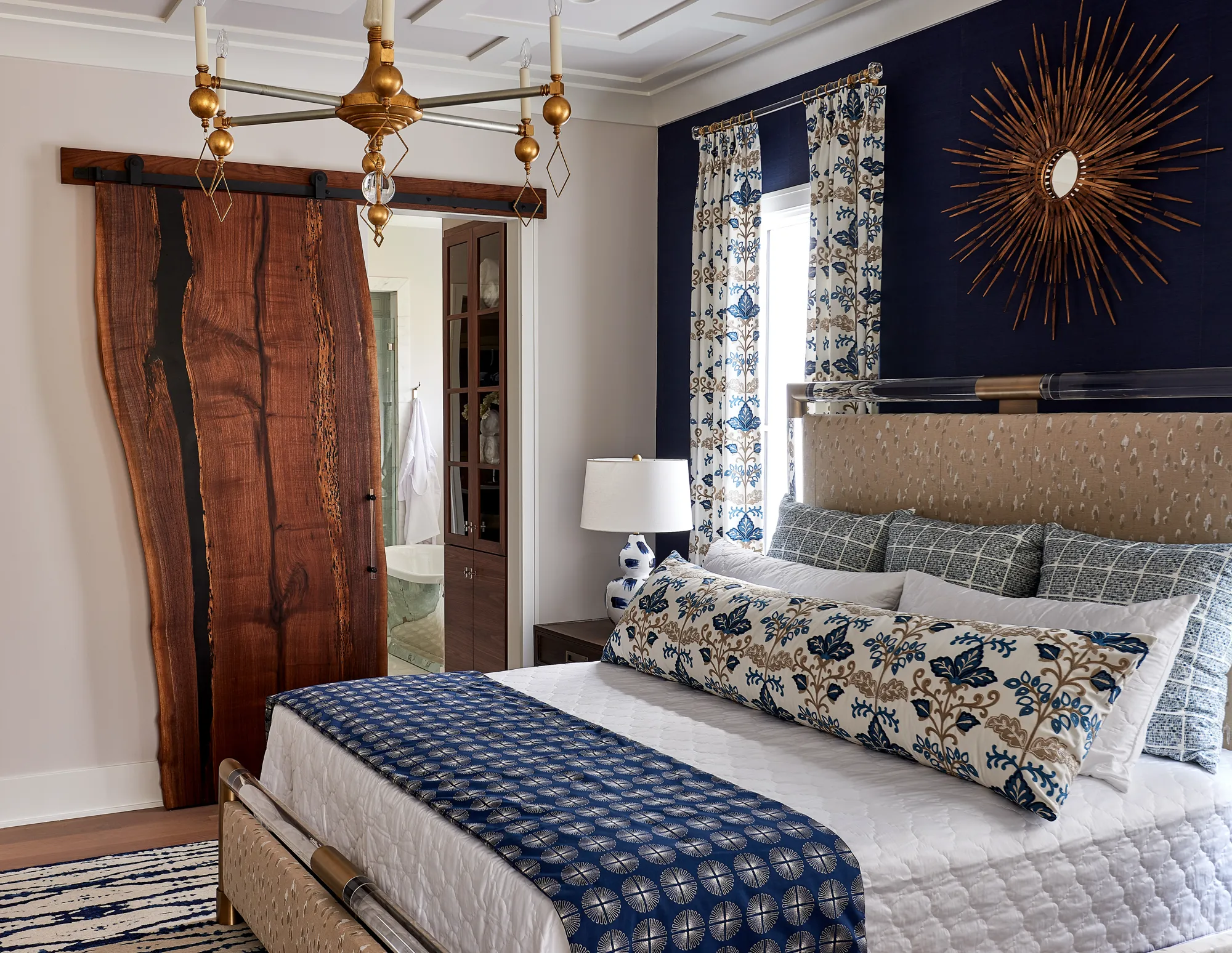 An elegant bedroom dominated by a large live-edge wood barn door and a dark blue accent wall with a gold sunburst mirror. A patterned bed is centered in the room.