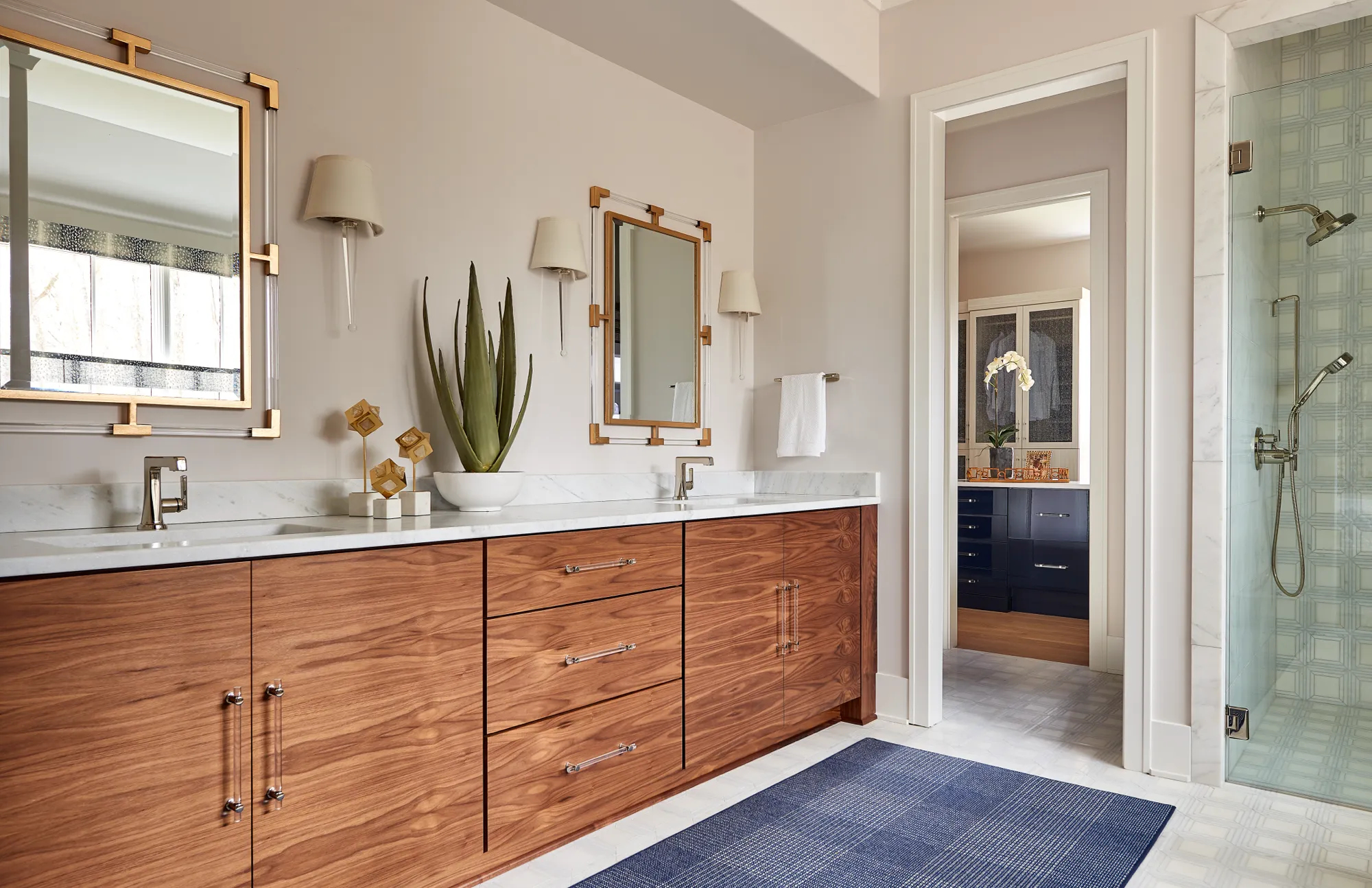 A modern, elegant bathroom with a rich wood dual vanity, marble counter, and polished gold accents. A blue rug sits on the white tile floor.