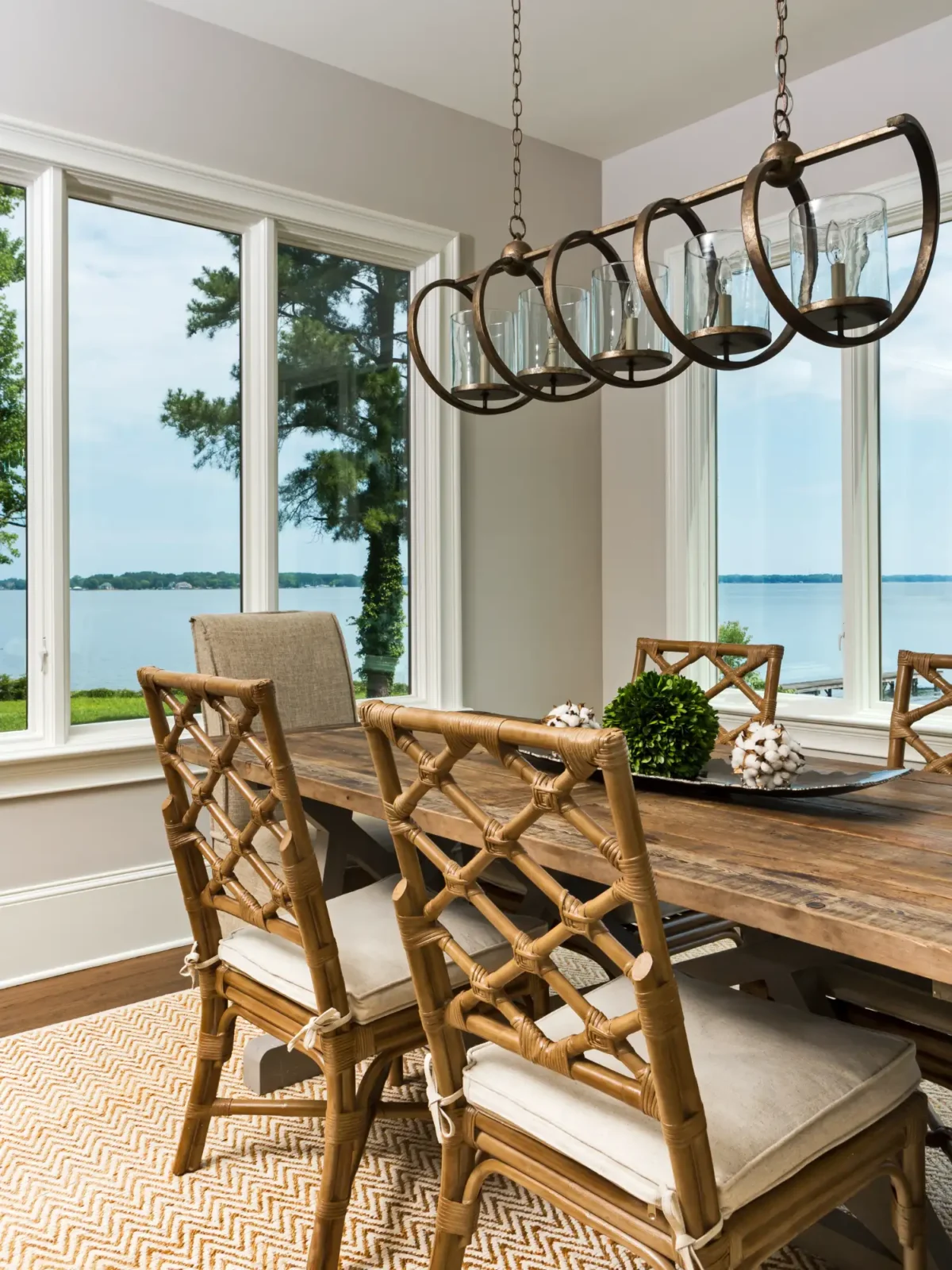 A rustic wooden dining table and bamboo chairs with linen cushions sit in a bright room overlooking a serene lake. A bronze linear chandelier hangs above the table.