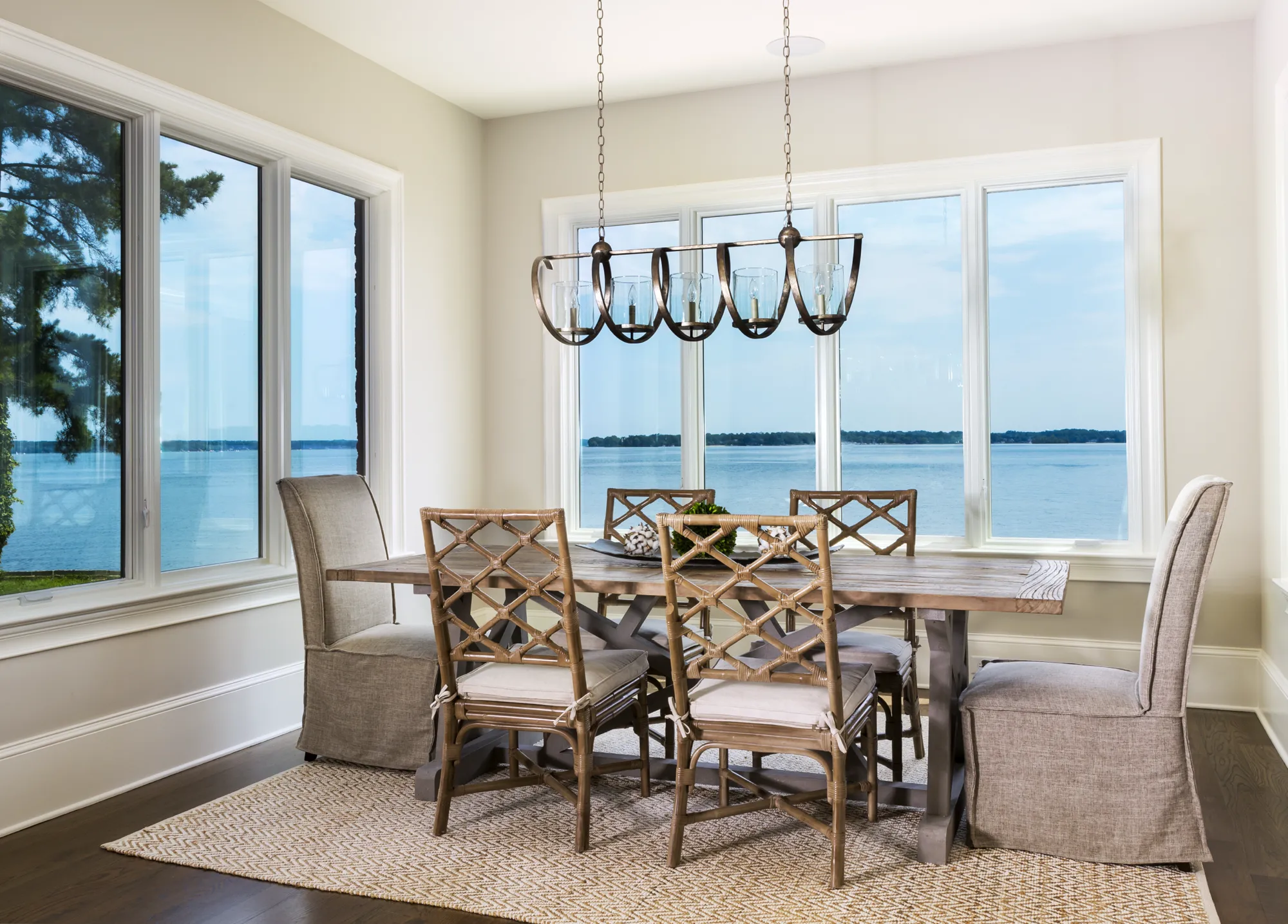 A bright dining room with a rustic wooden table and mixed seating, set against expansive windows overlooking a calm, blue water. A metal chandelier lights the scene.