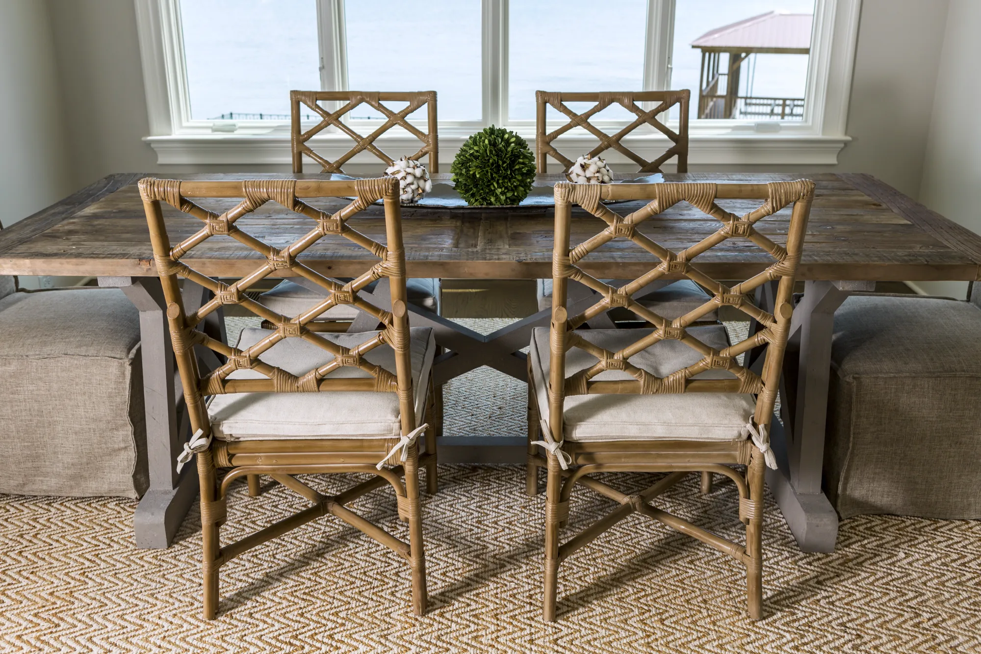 A coastal dining room features a rustic wood table, four rattan chairs with cream cushions, and a chevron rug. The centerpiece includes a green topiary, set before large bright windows.
