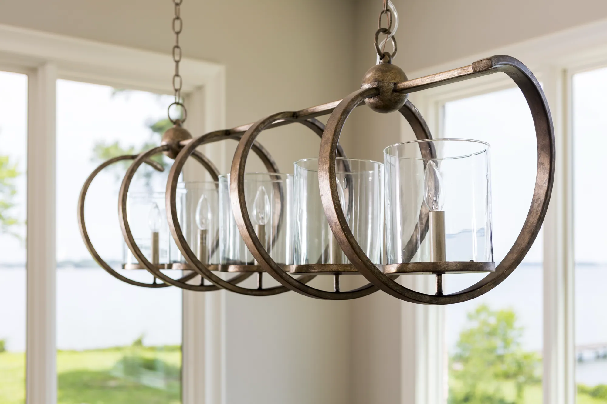 A linear bronze chandelier hangs, featuring four bulbs enclosed in clear glass cylinders. The background shows large windows overlooking a waterfront landscape.