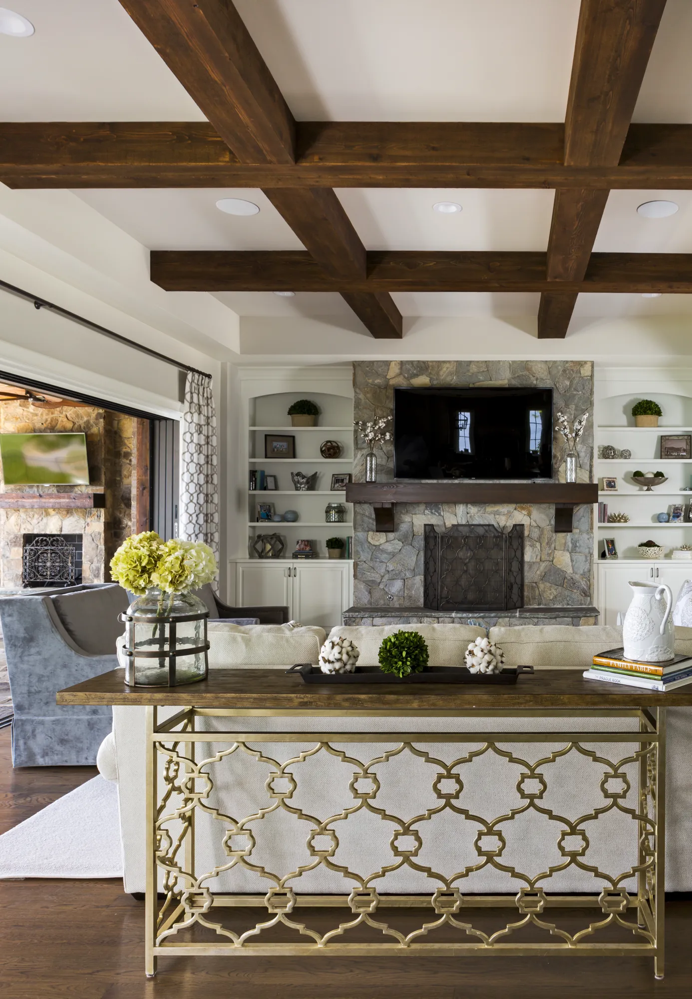 A rustic-chic living room with dark wood ceiling beams and a large stone fireplace. A gold console table displays flowers, cotton decor, and books in the foreground.