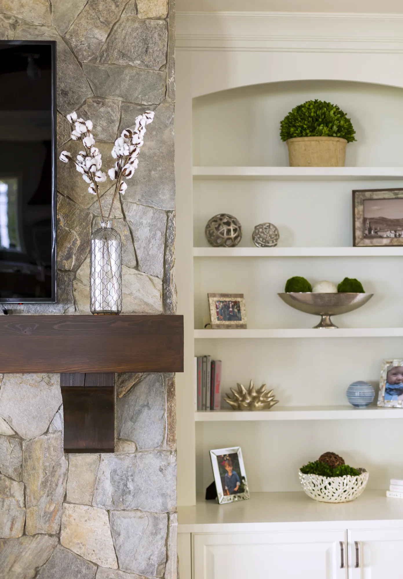 A dark wood mantle on a stone fireplace holds cotton stalks in a vase, next to white built-in shelving displaying framed photos and décor.