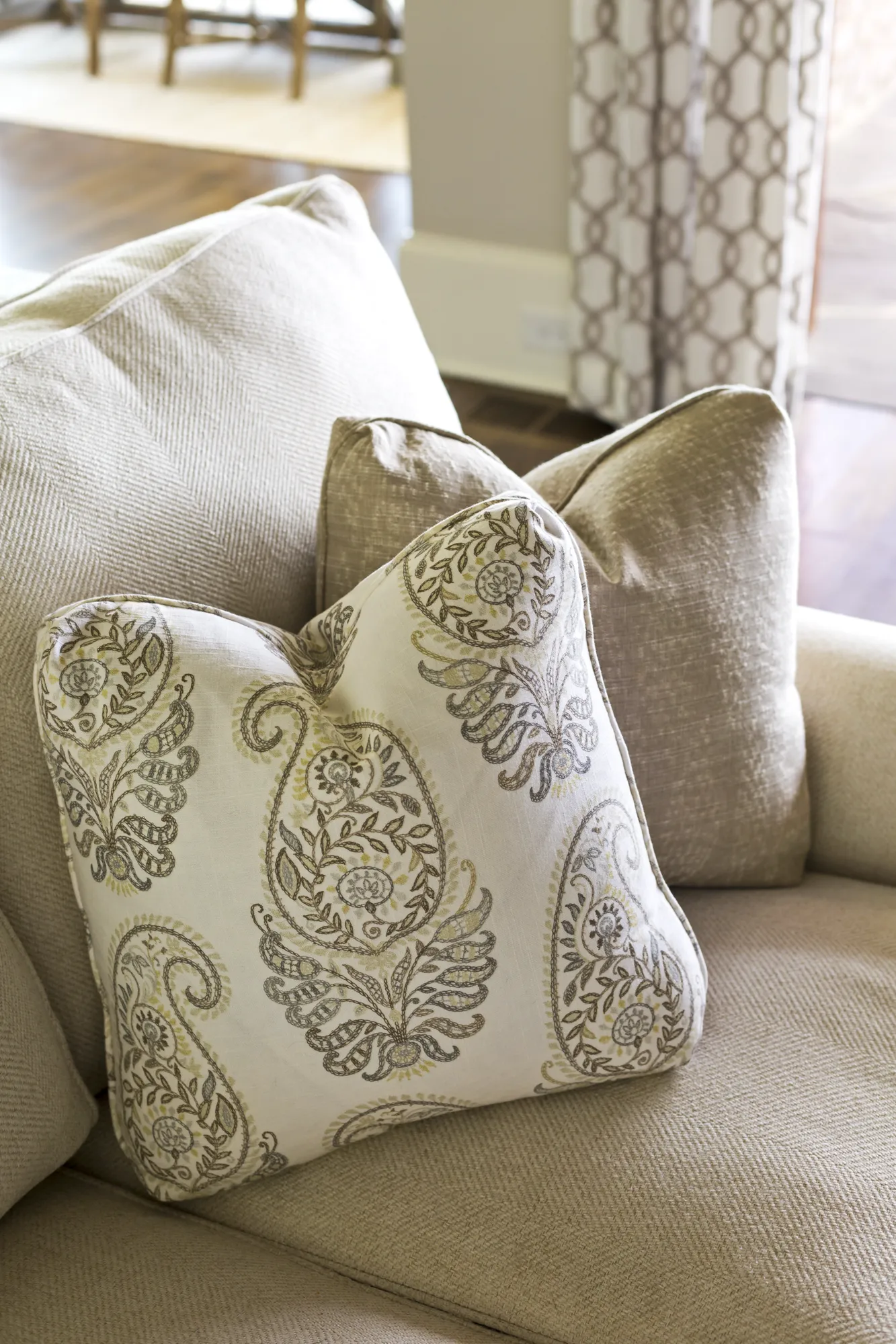 A decorative pillow featuring an ornate, muted paisley pattern rests against solid neutral cushions on a textured sofa.