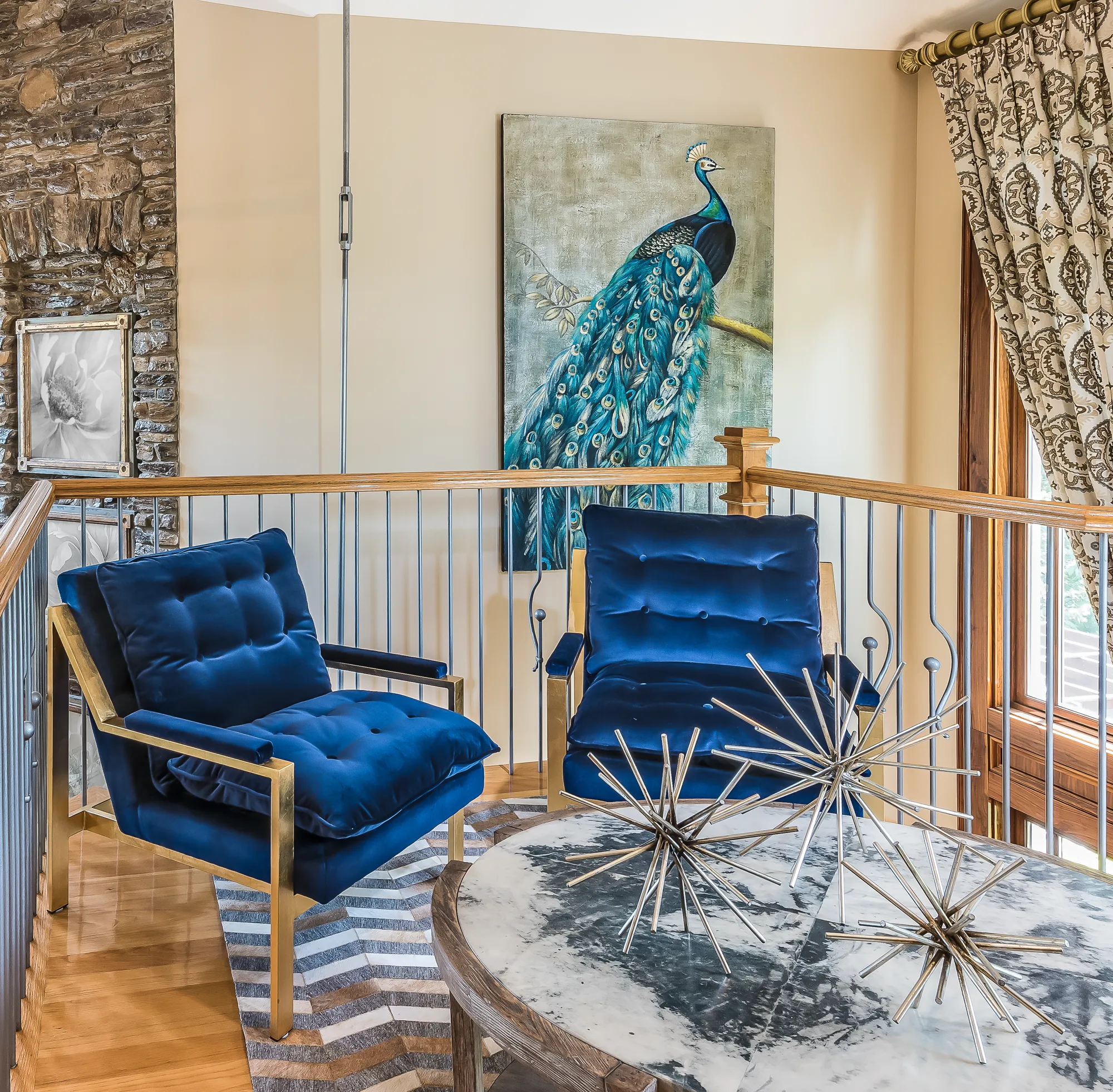 Two luxurious navy blue velvet armchairs with brass frames sit on a loft landing next to a round marble table and a large peacock painting.