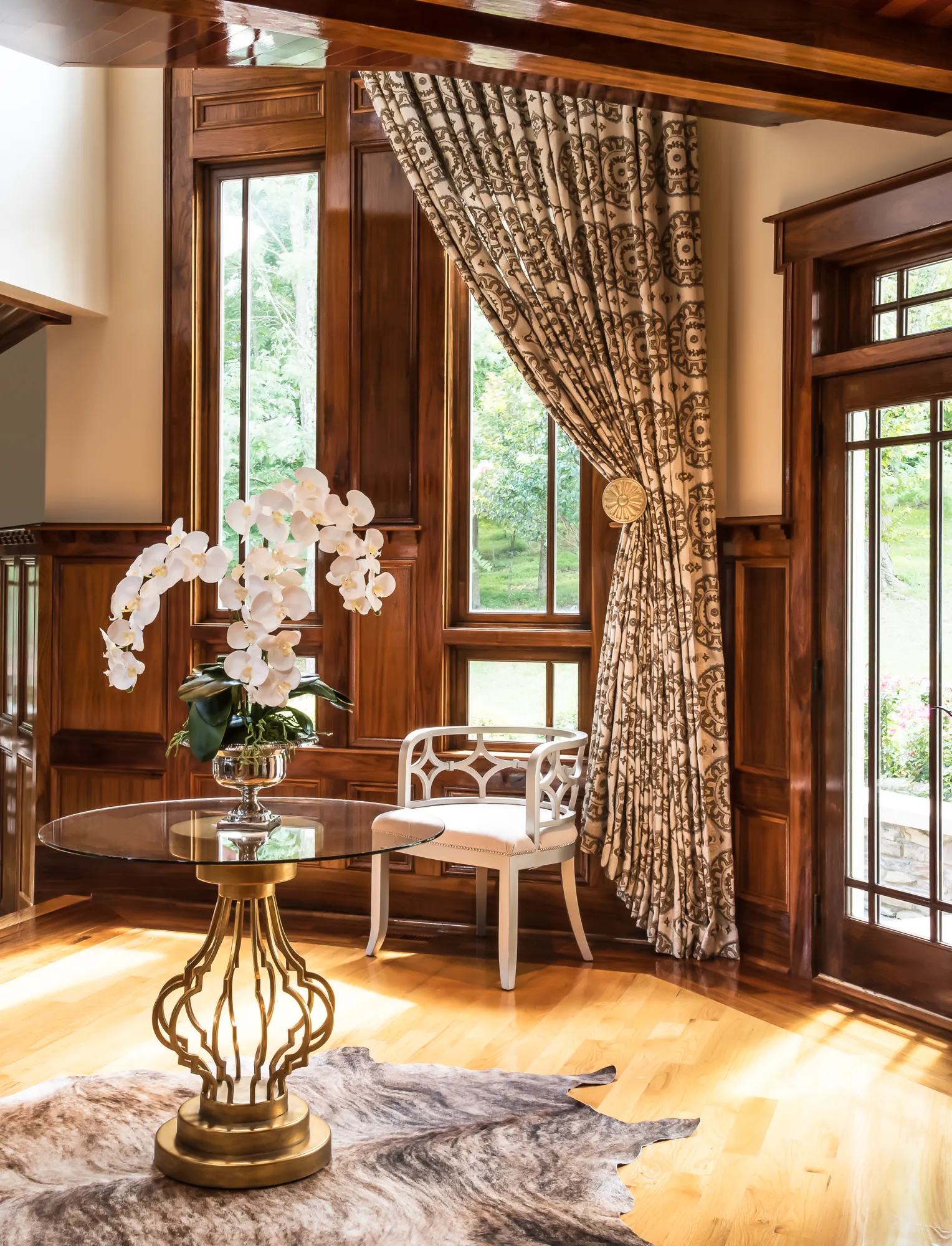 An elegant interior corner with dark wood paneling and wainscoting features a gold-base glass table holding white orchids, a decorative white chair, and a patterned curtain.