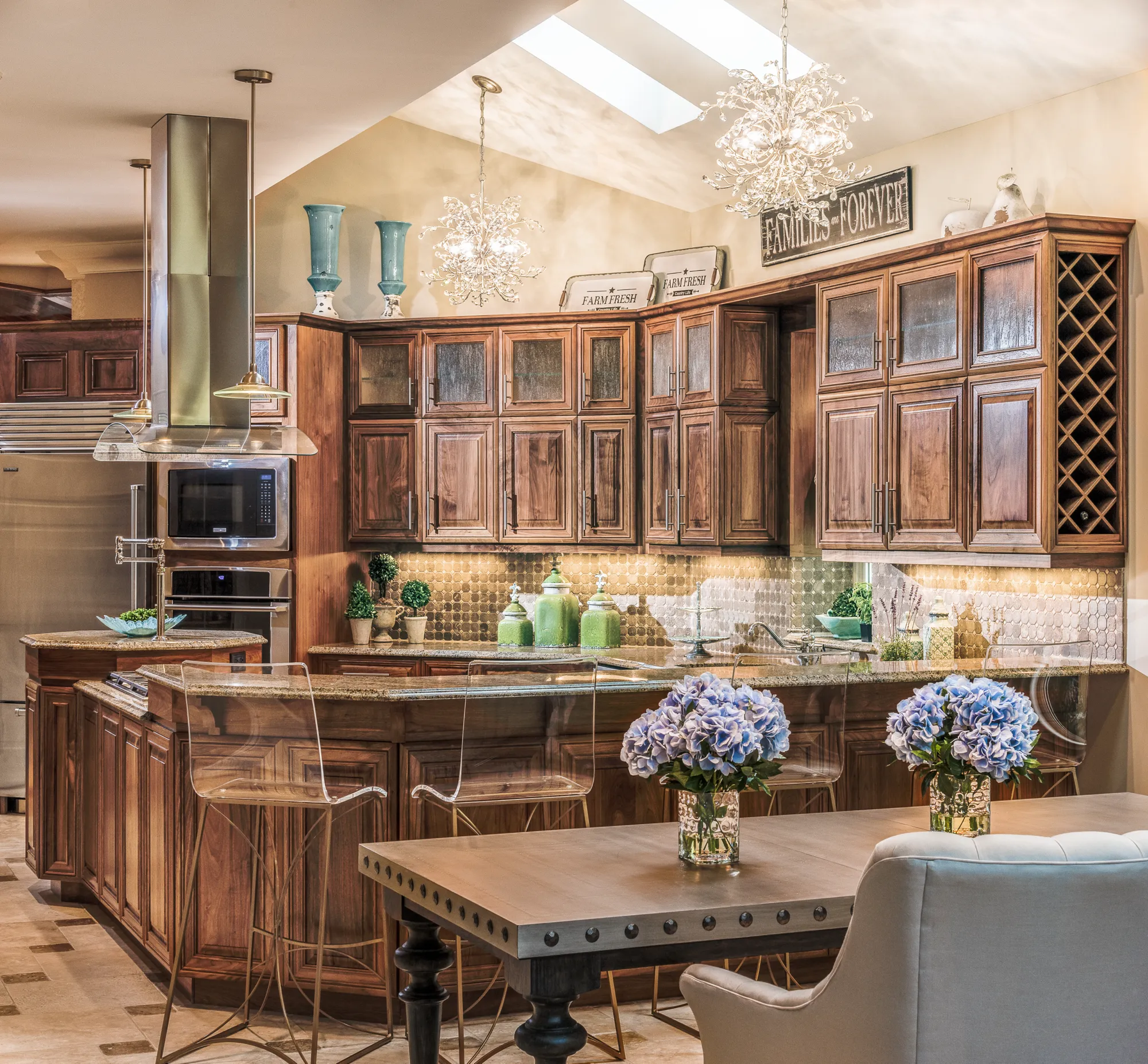 A luxurious kitchen featuring dark wood cabinets, granite counters, stainless steel appliances, and bright under-cabinet lighting. Crystal chandeliers and hydrangeas add elegance.